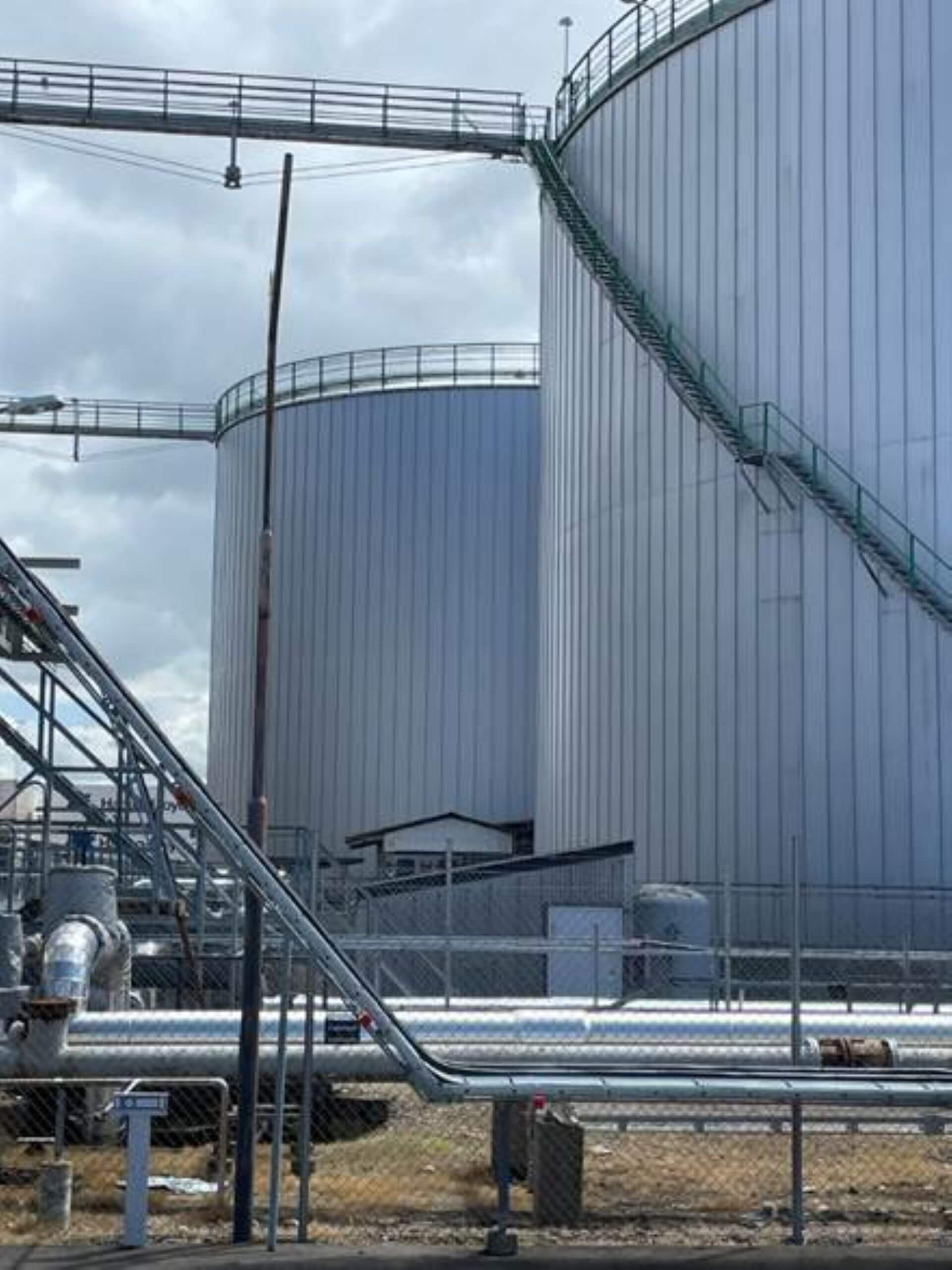 Close up of wire fend with large cylindrical storage tanks behind and pipework running left to right across photo