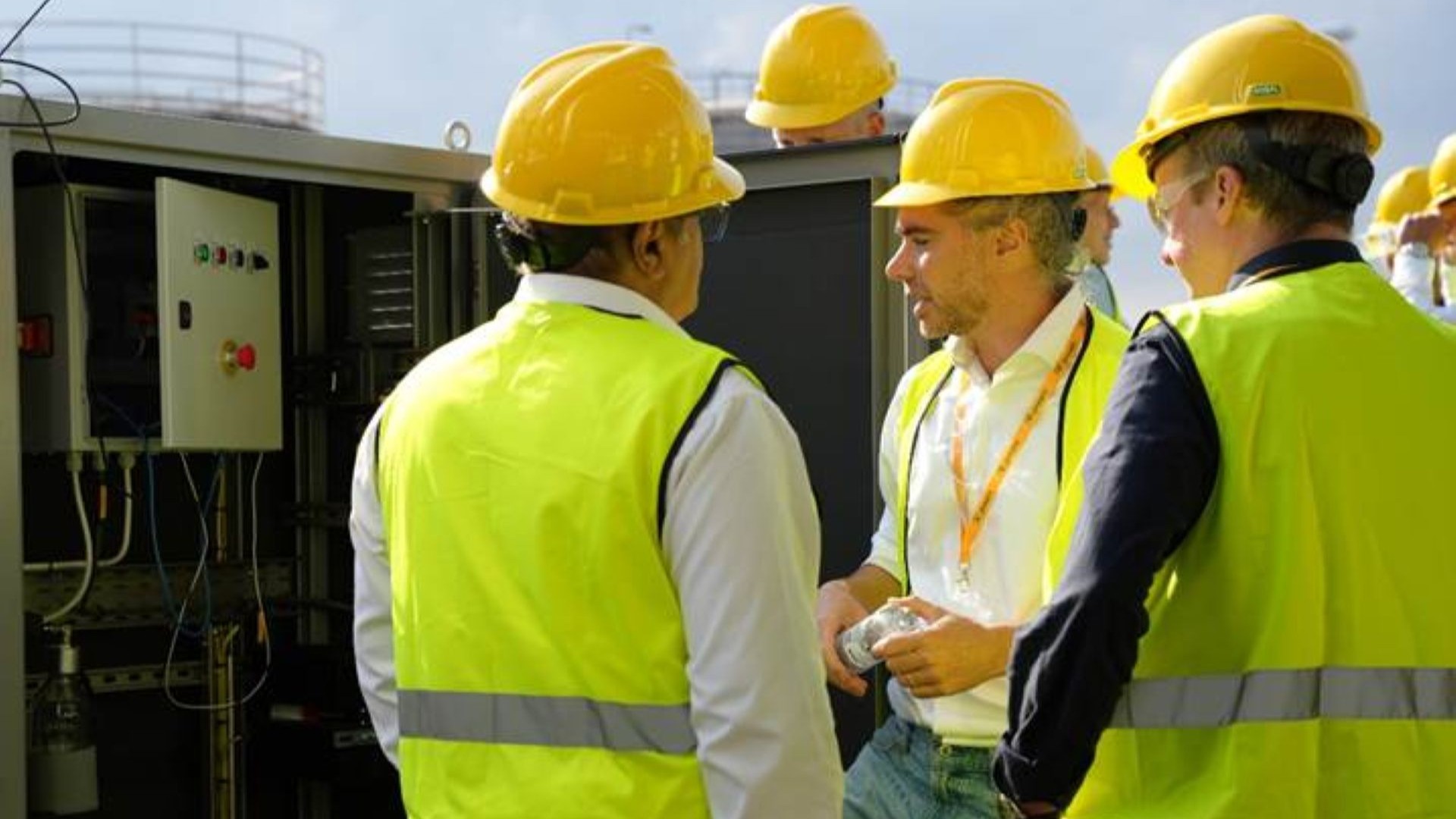 Three men in high-viz jackets and hard hats chatting in front of opened control box, tops of bulk storage tanks just visible in background