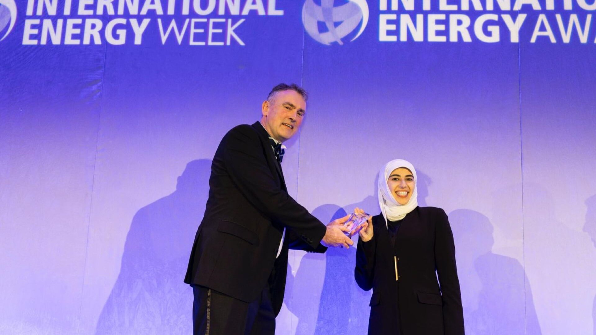 Man presenting award to woman, on stage, with International Energy Week logo projected on screen behind