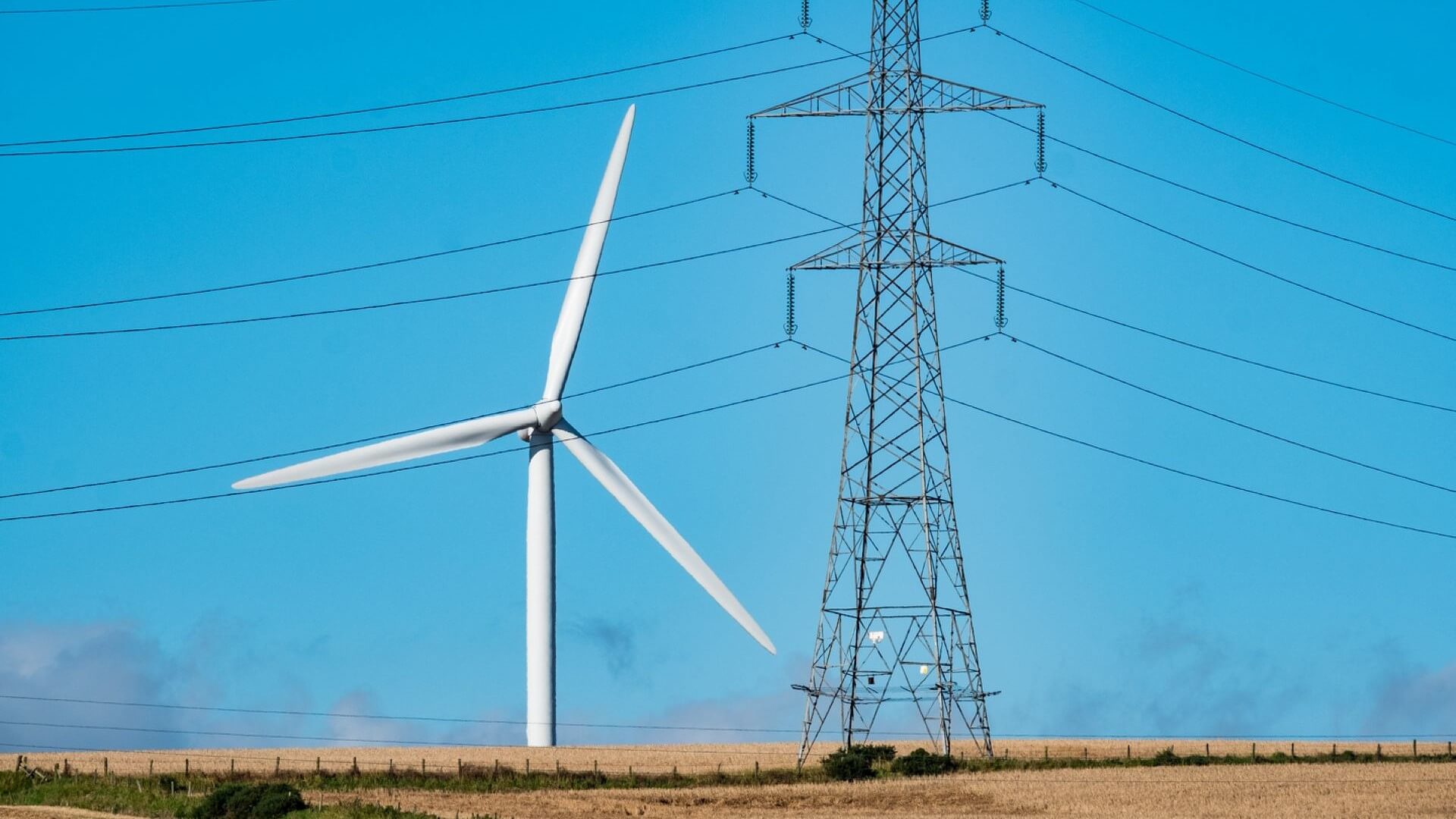 Wind turbine close to an electricity pylon, with blue sky behind