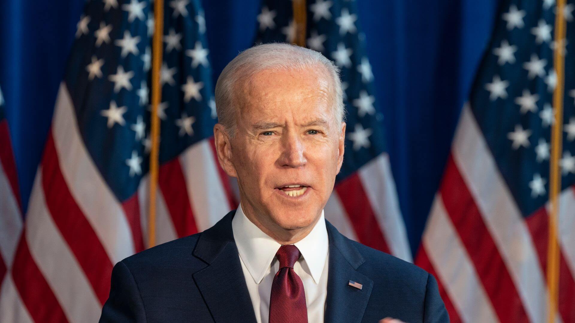 Head and shoulders image of US President Joe Biden standing at lecturn with US flags set behind him