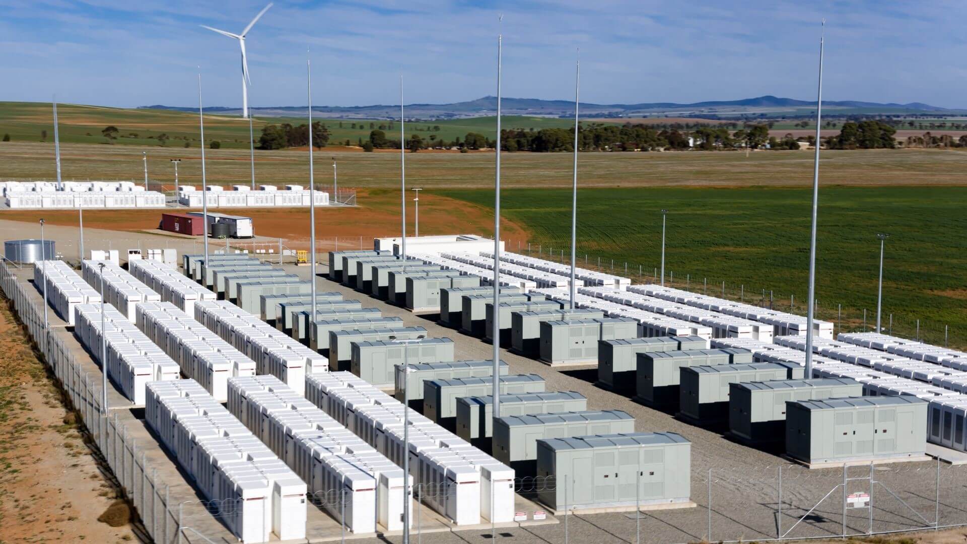 Aerial view over Hornsdale Power Reserve showing rows of battery units in foreground and wind tubines in distance