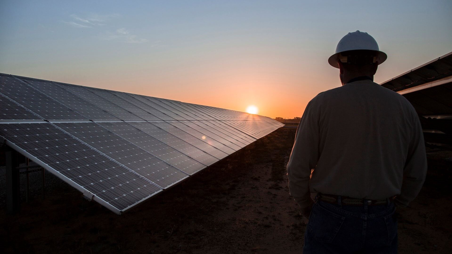 Engineer wearing hard hat, standing by solar panels in solar farm, set against orange sunrise sky