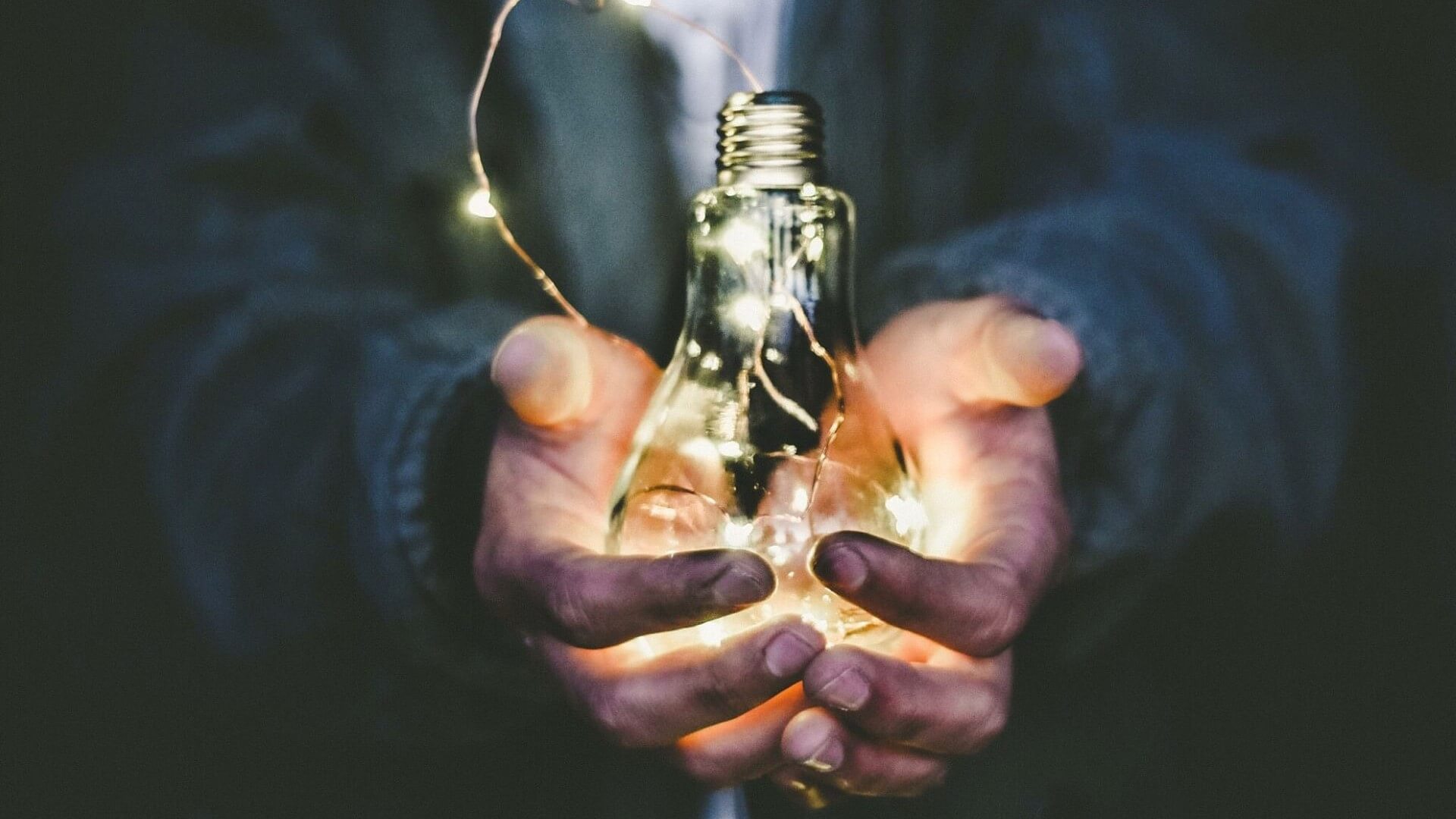 Man in dark suit, set against dark background, cupping lighted lightbulb in hands
