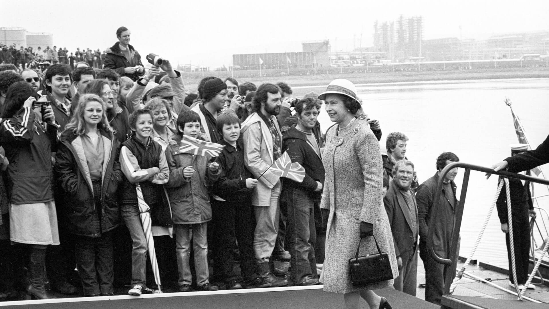 Queen walking in front of cheering crowds, with Sullom Voe terminal in the distant background