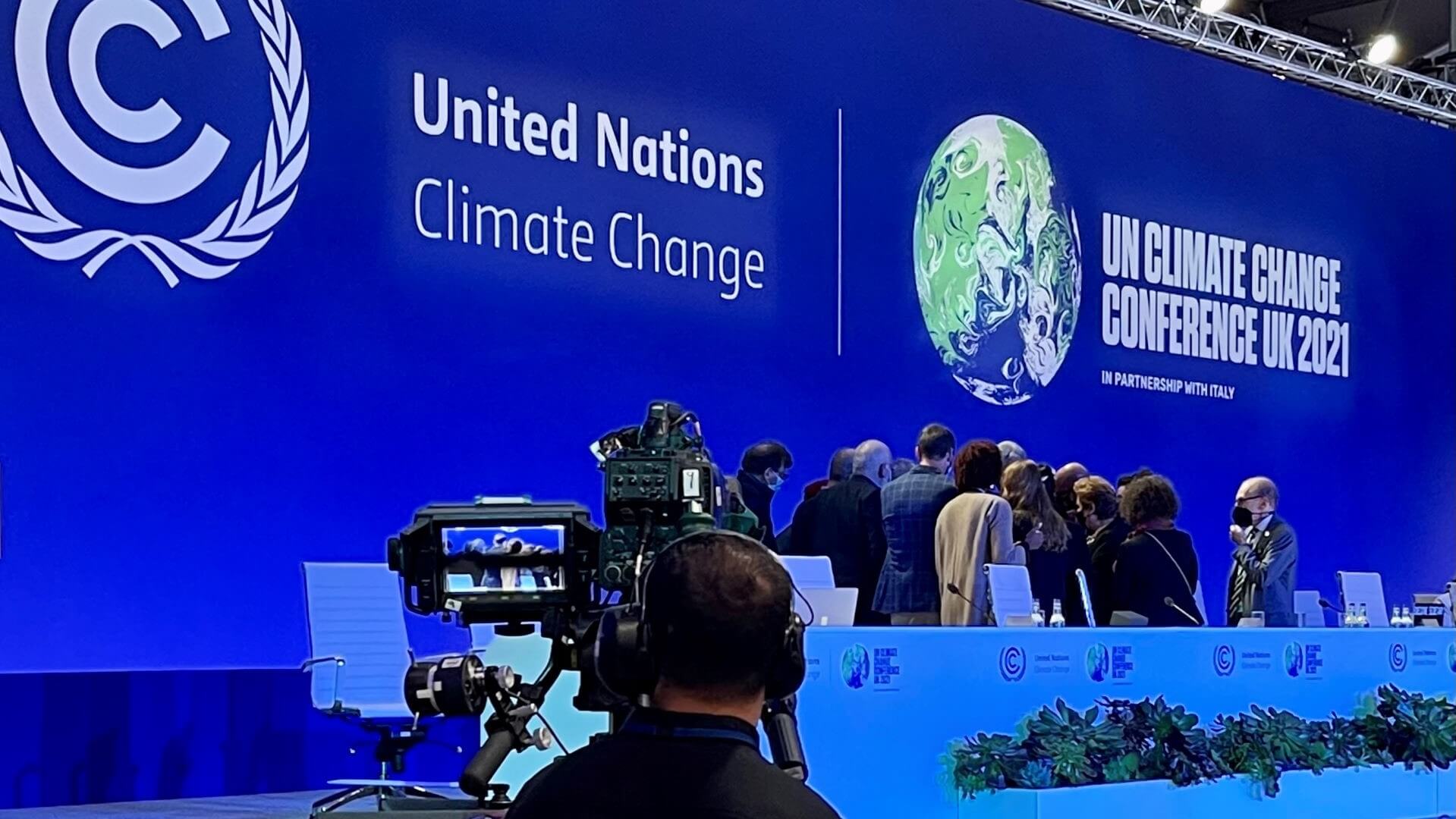 Huddle of speakers by top table at COP26 plenary session with TV camera and cameraman in foreground