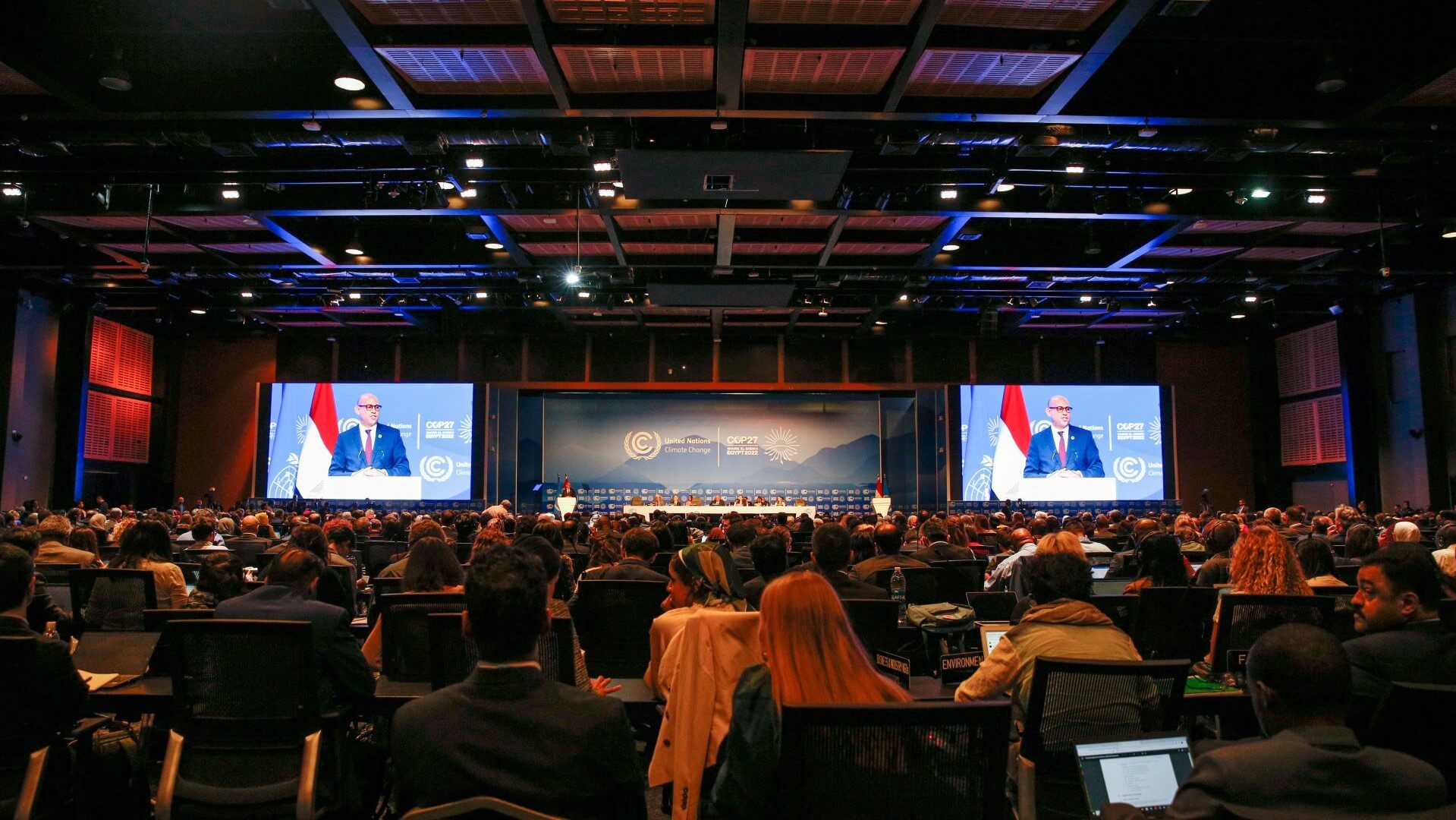 View of packed audience listing to COP27 address from podium and shown on two large TV screens