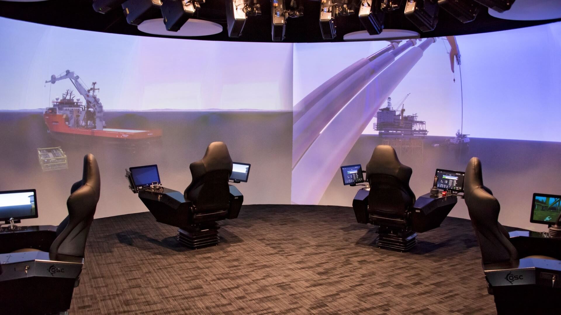 Simulator room with large chairs with computer screens on each arm, with large projection screens in background