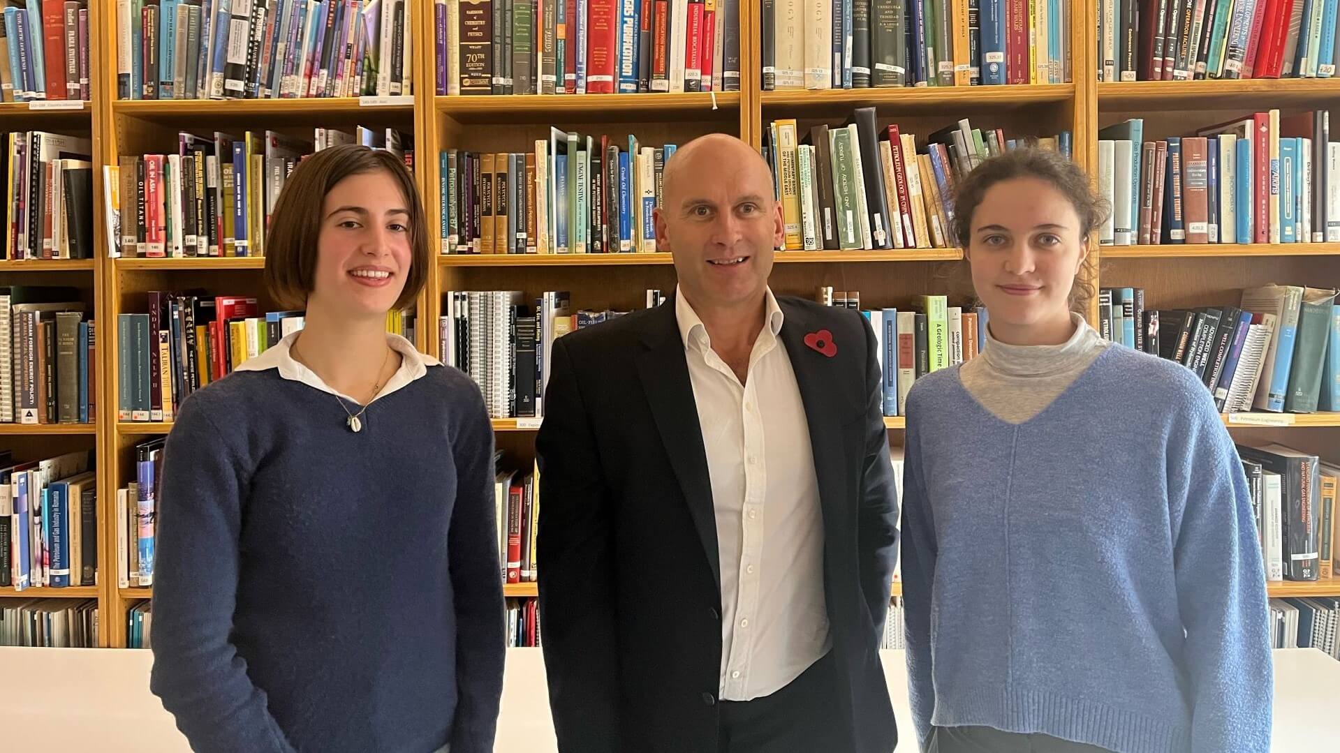 Two students standing either side of EI CEO, with library shelving full of books behind