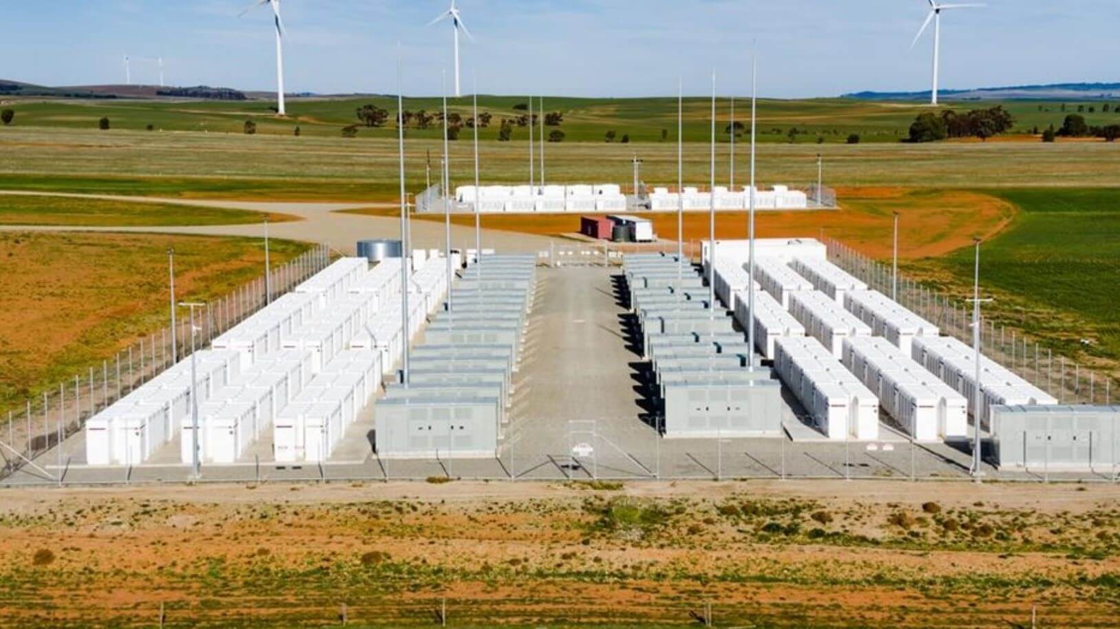 Aerial view over Hornsdale Power Reserve showing rows of battery units in foreground and wind tubines in distance