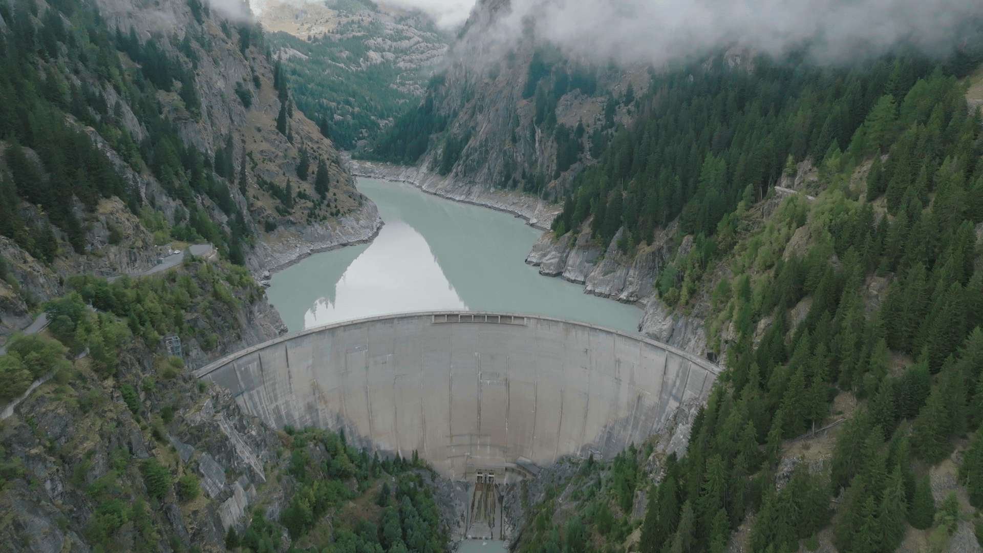 Aerial view over hydropower dam, looking at front of dam wall with water behind and forests on mountain sides either side of dam