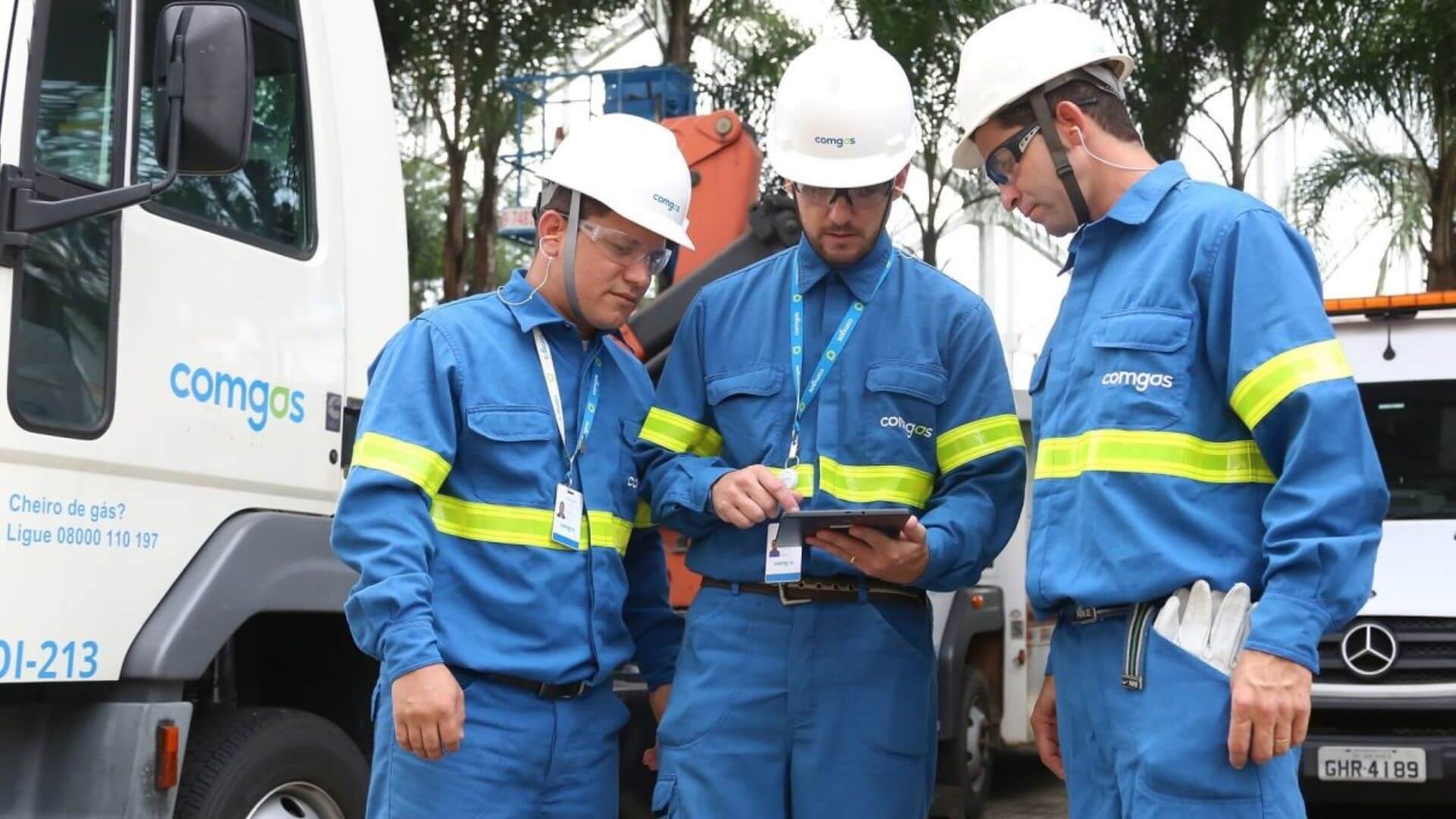 Three Comgás engineers in company's blue overalls and white hard hats looking at a tablet while standing in front of a truck
