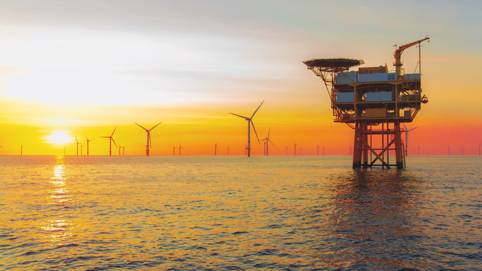 Offshore oil and gas platform in foreground with wind farm in background and set against orange sunset sky
