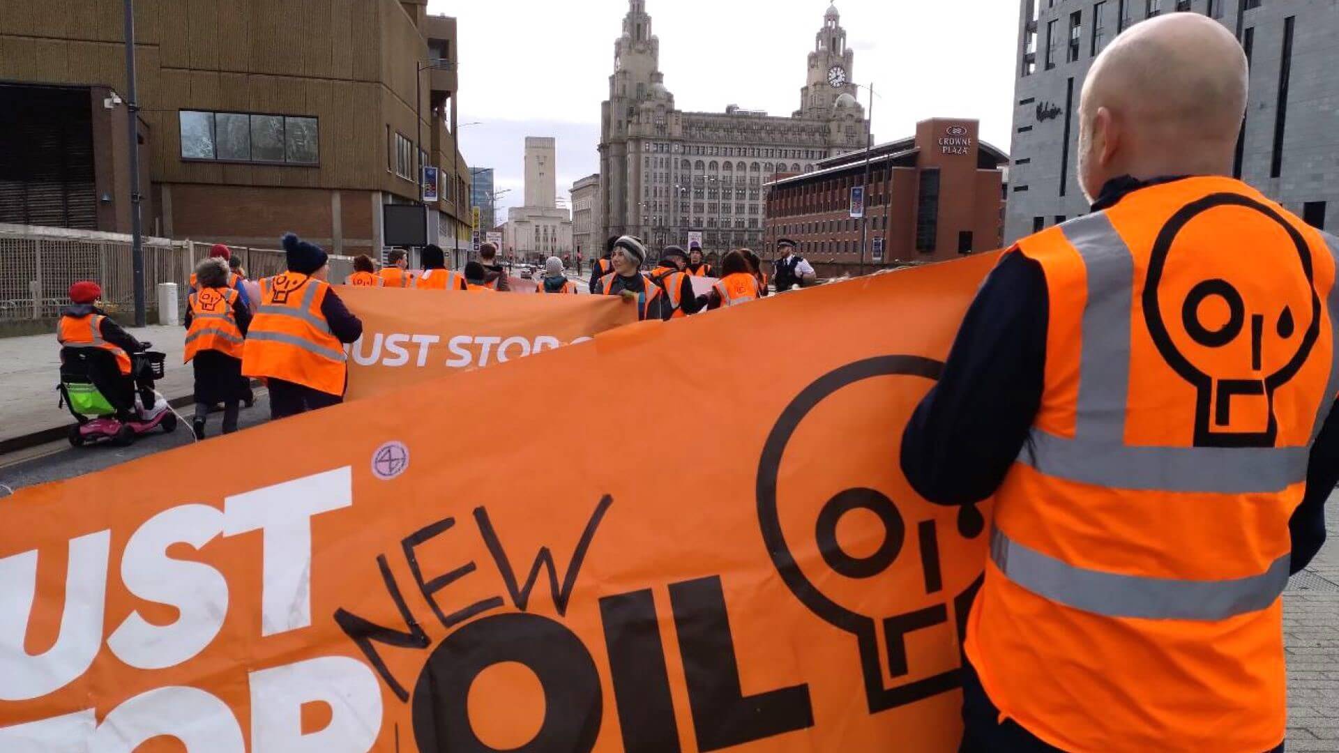 Close up of Just Stop Oil orange banner being held by a protester with back to camera and other protesters in orange jackets extending into distance
