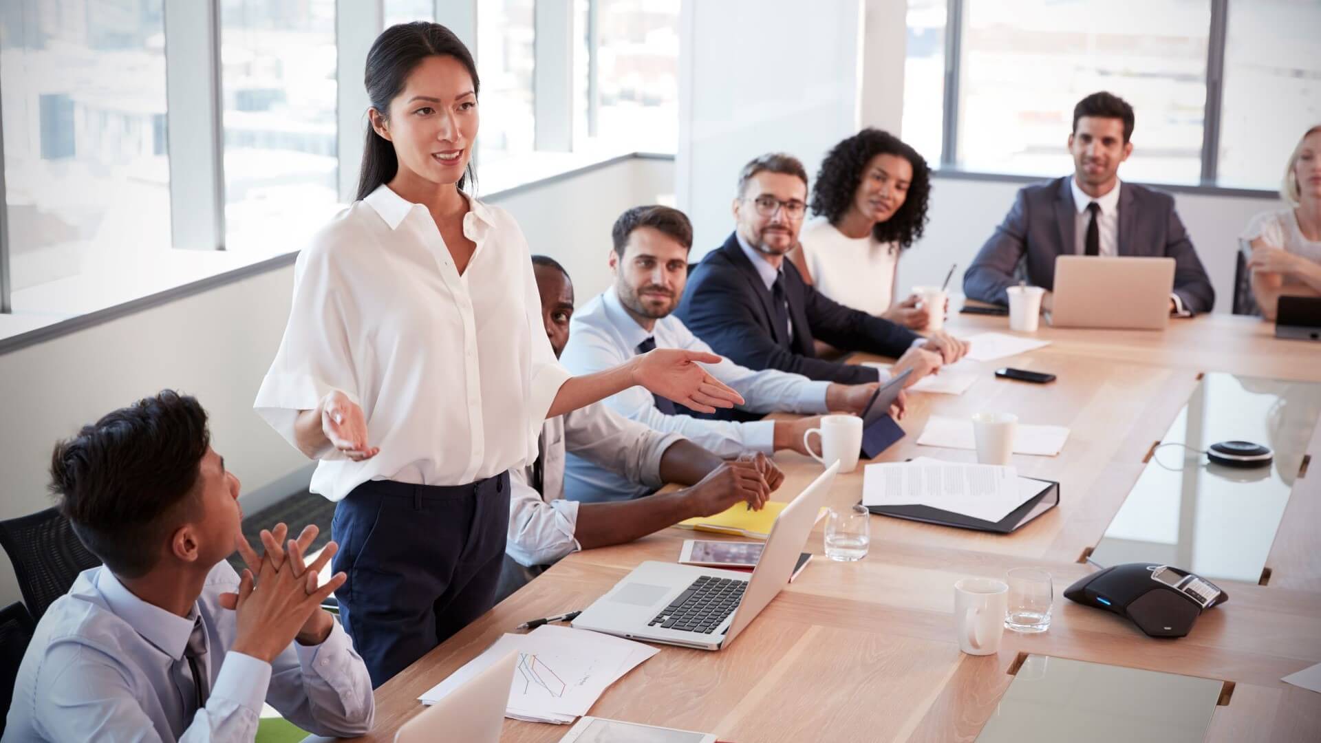 Business people of different ethnicity, age and gender sitting around table with one woman standing up and making a presentation