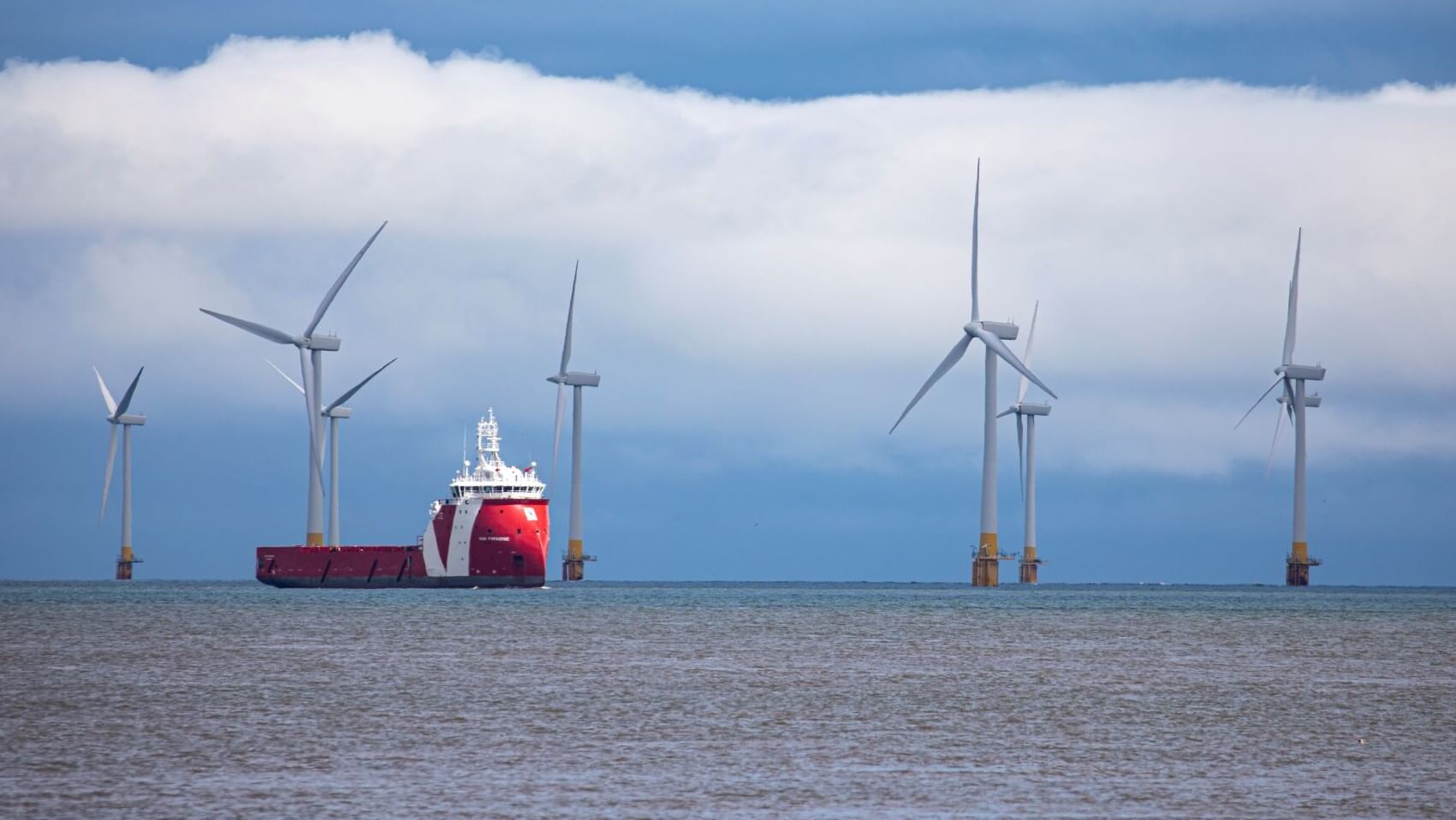 Seven offshore wind turbines and a vessel at sea, set against cloudy sky