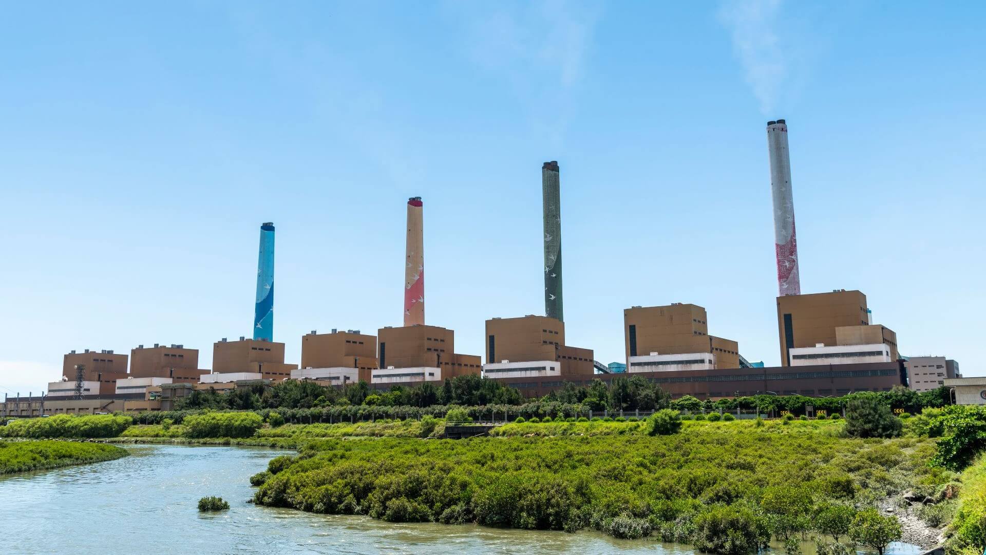 Row of coal plant and chimney set against blue sky, with water and green vegetation in foreground