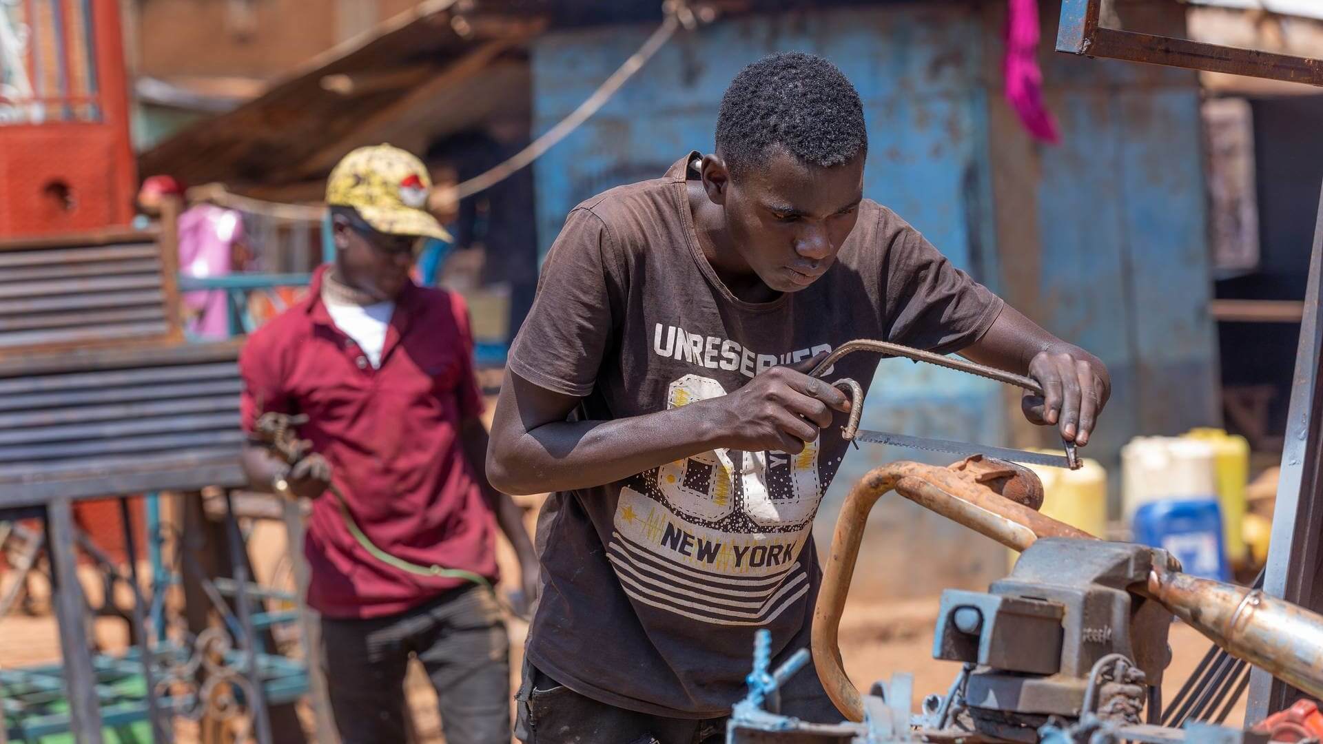African man working outside, using a hacksaw on pipework clamped in vice