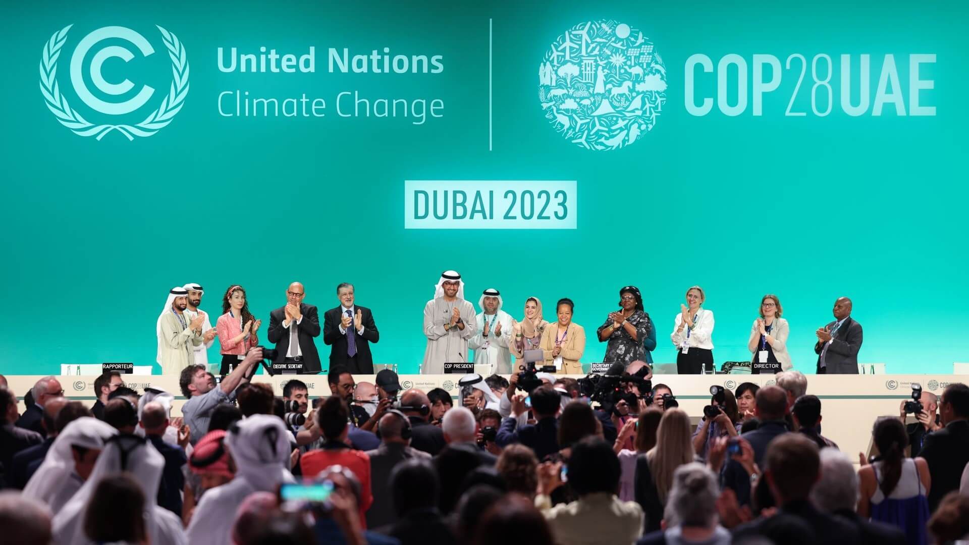 Men and women standing behind desk on stage, clapping hands, in front of audience of people standing up
