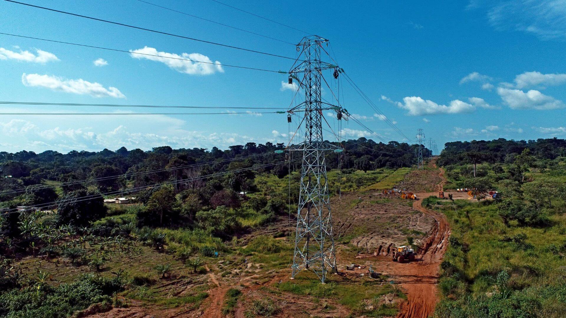 Electricity pylon and transmission wires, standing amidst green trees to left and right and construction work taking place underneath in red orange soil