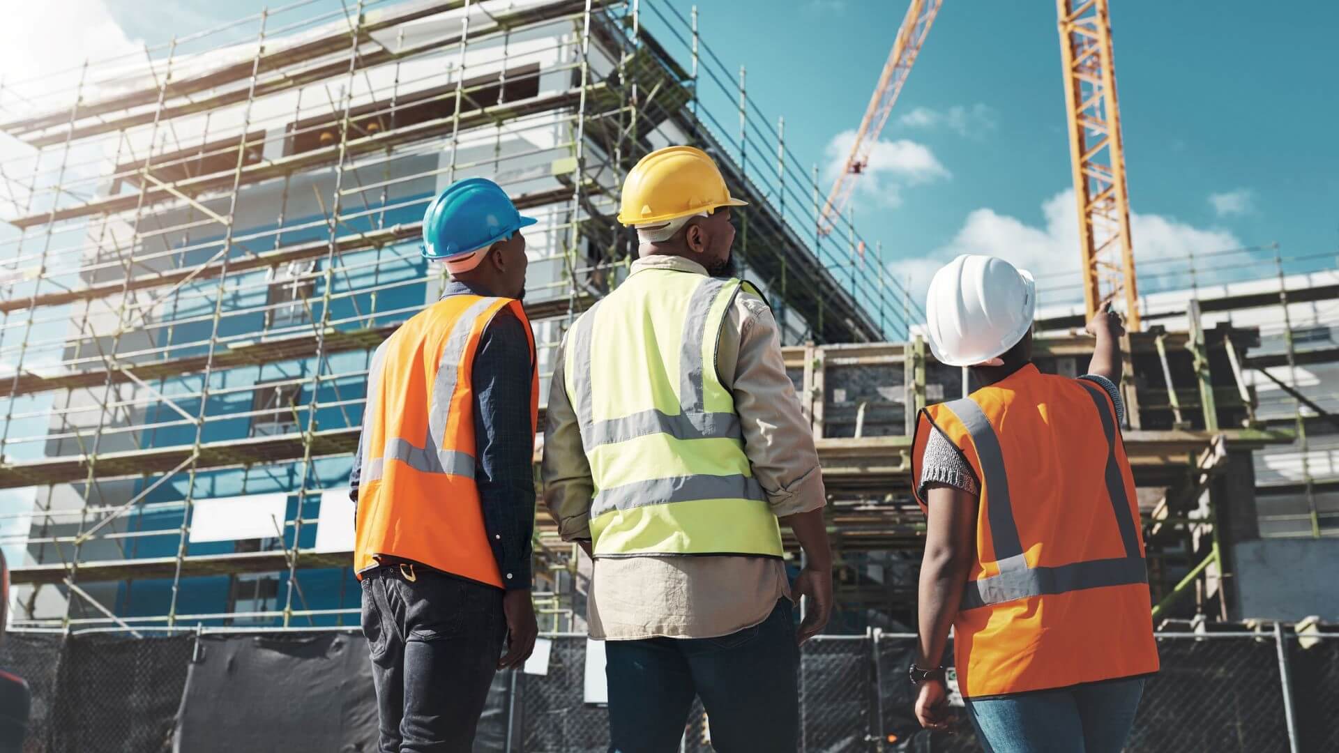 Three construction workers in hard hats and hi-viz jackets looking at crane and building under construction 