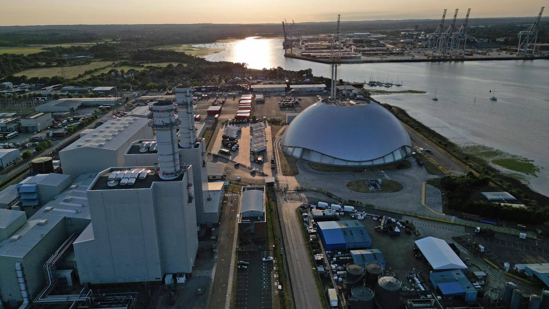Aerial view over gas-fired power station facilities at sunset, with container terminal on horizon