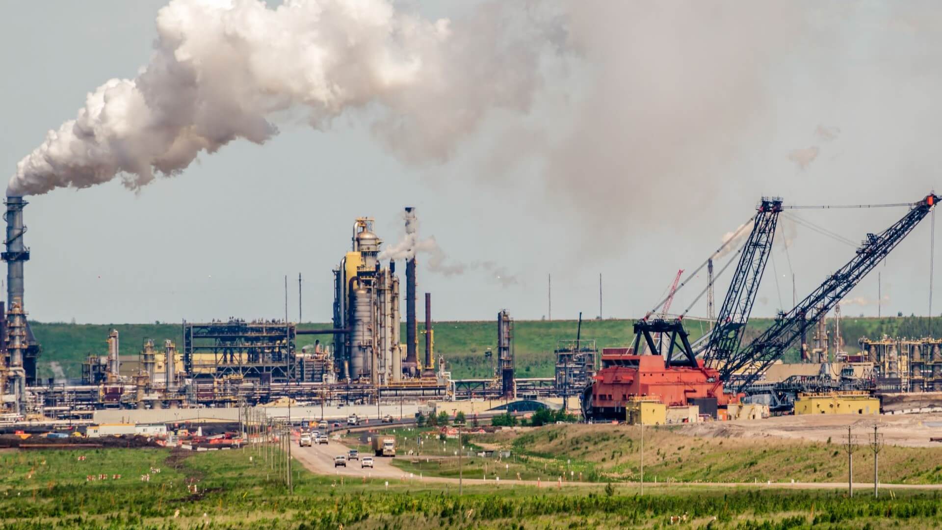Distant view of crane and machinery working in industrial oil sands, with smoking chimney stacks to left