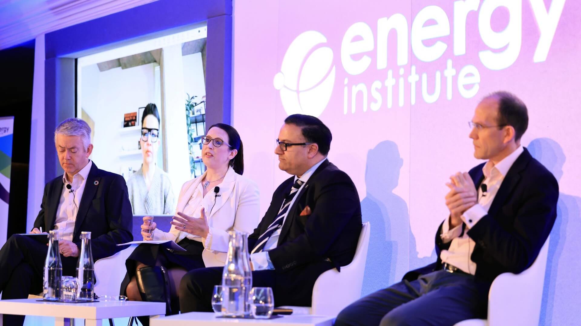 Three men and one women in panel discussion, sat on stage with screen projection behind their seats