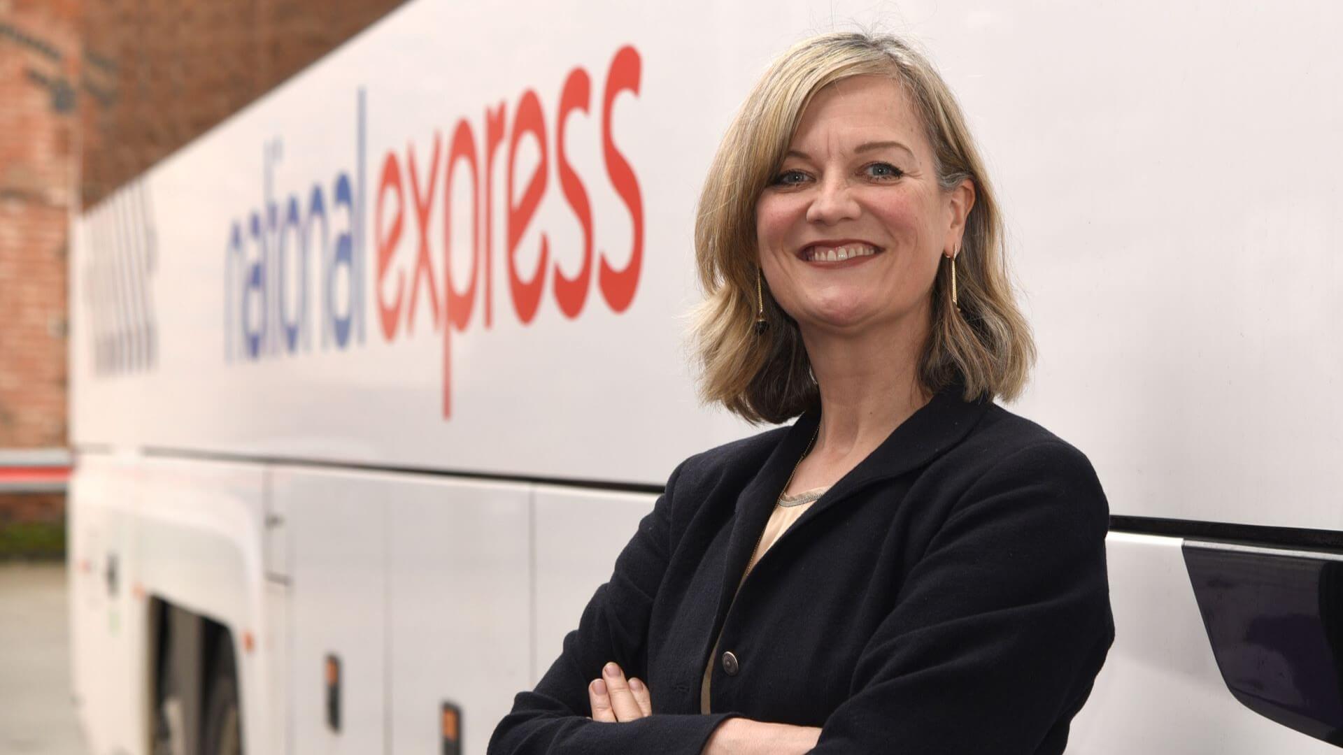 Head and shoulders shot of Alex Jensen, CEO, National Express, standing with her arms crossed alongside coach with company log