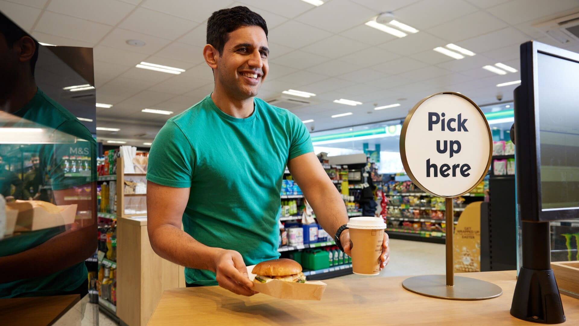 Man at a forecourt cafe passing a burger and take away cup of coffee across the counter to a customer
