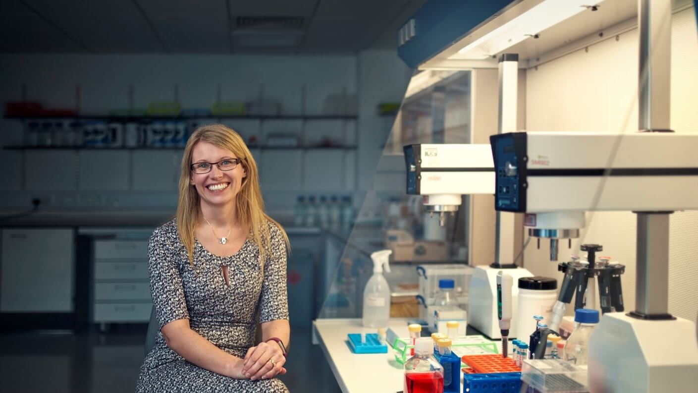 Photo of Professor Aline Miller, facing the camera, sitting alongisde a laboratory bench with microscopes, flasks, vials and other lab equipment