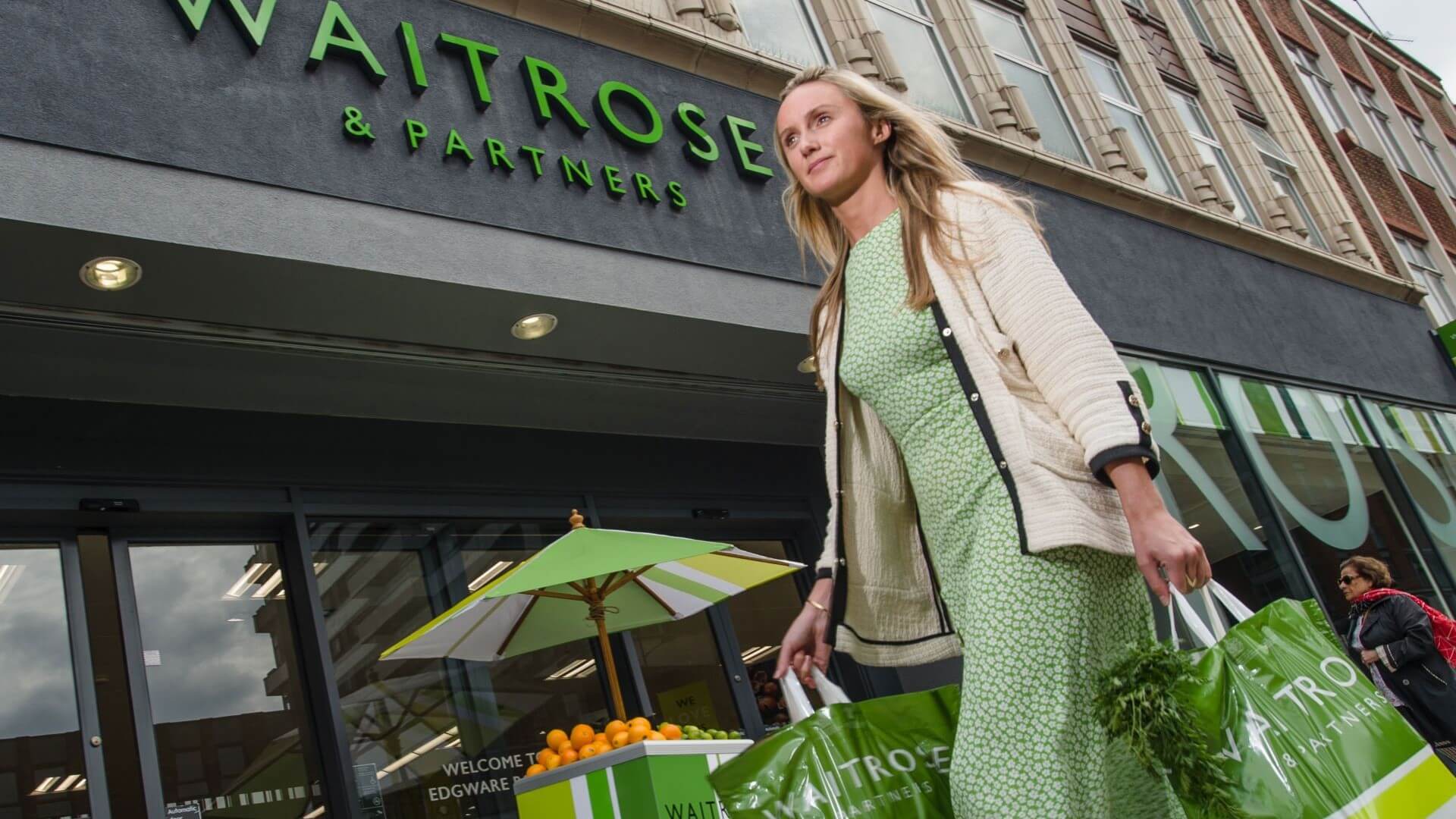 Woman carrying plastic shopping bags walking past the front of a Waitrose supermarket 