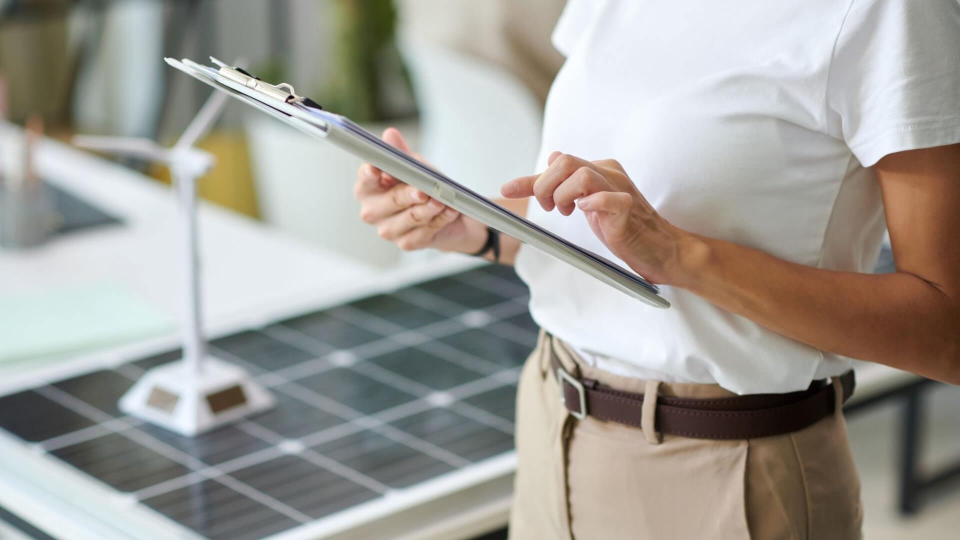 Close up of man holding computer tablet and typing with one finger, with solar panel and wind turbine models on desk alongside