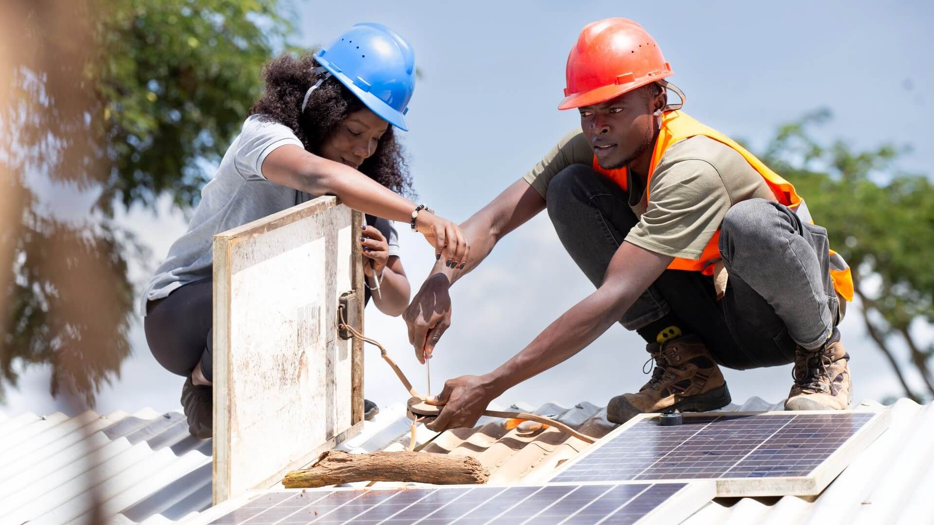 Woman in blue hard hat squatting on rooftop helping a man in a red hard hat install a solar panel