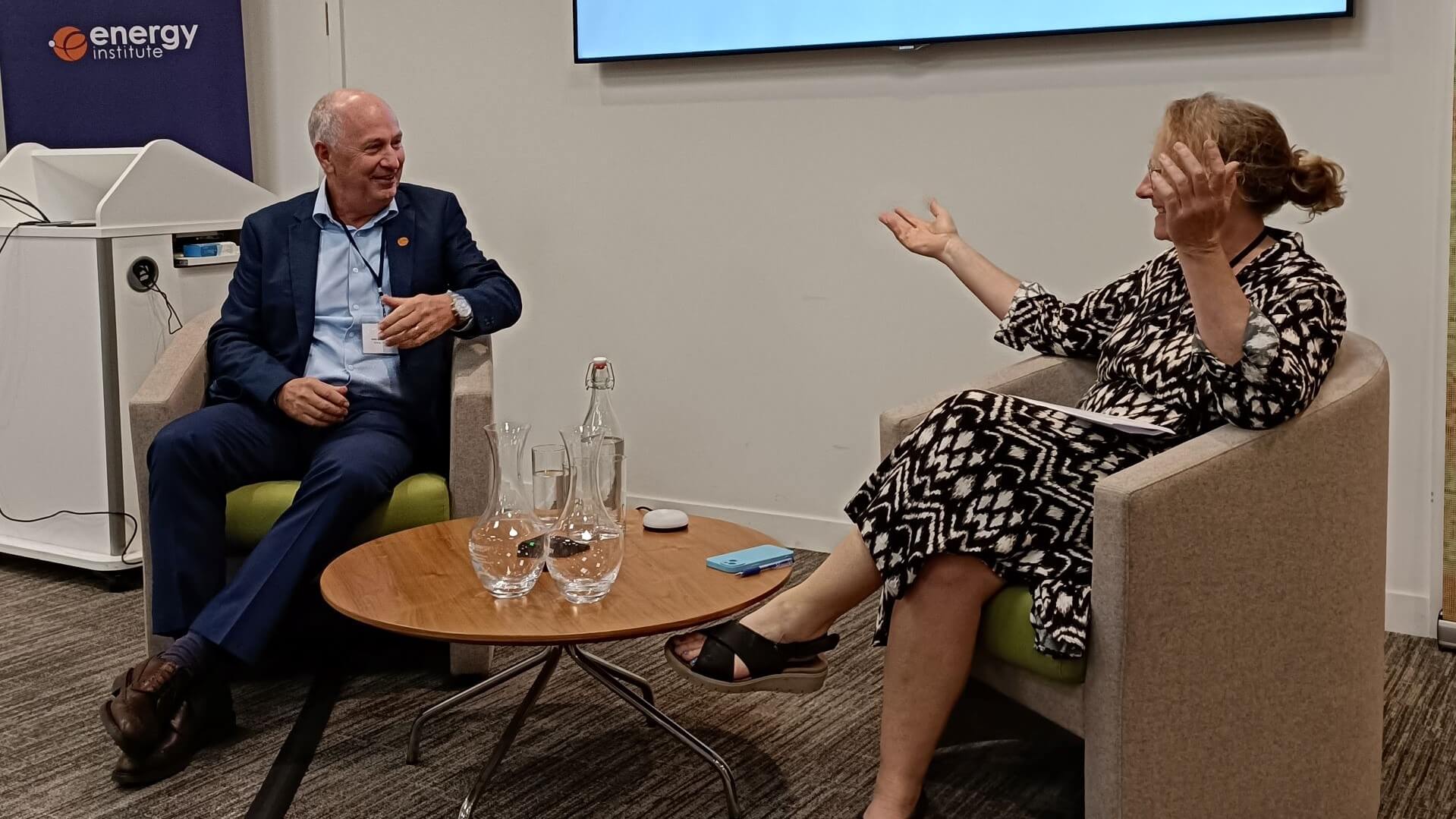 Man seated on stage to left of picture, chatting to woman sat to right, both smiling
