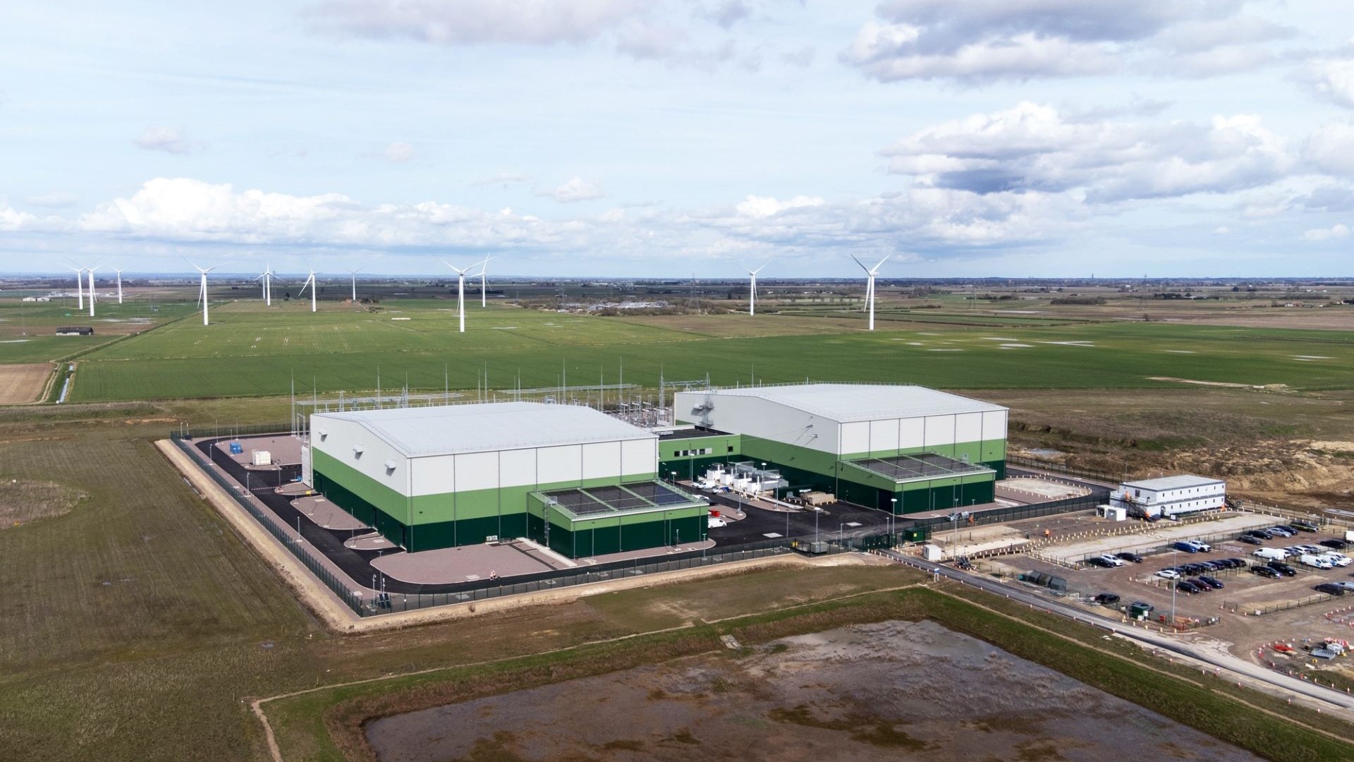 Aerial view over two large industrial buildings with car park in front, and green fields to rear, with wind turbines on distant horizon