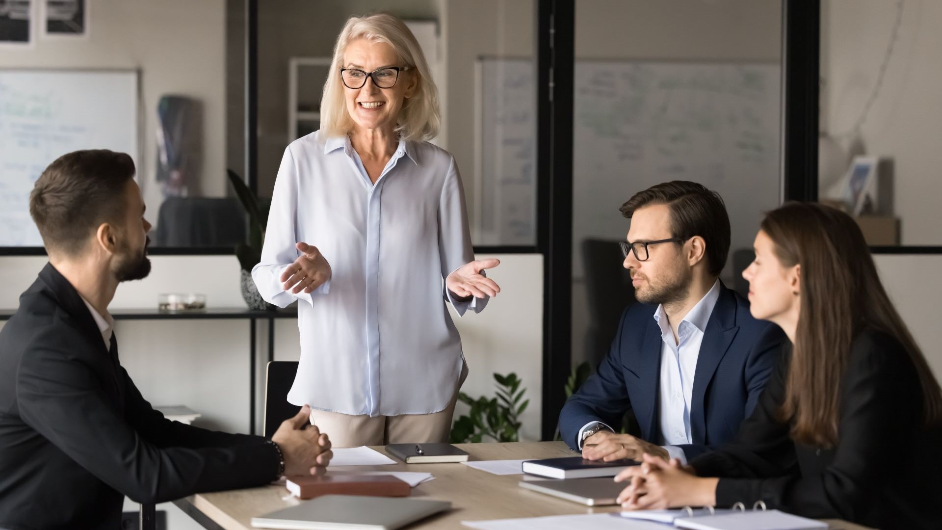 Two younger men and one woman sat at an office table listening to an older woman stood between them