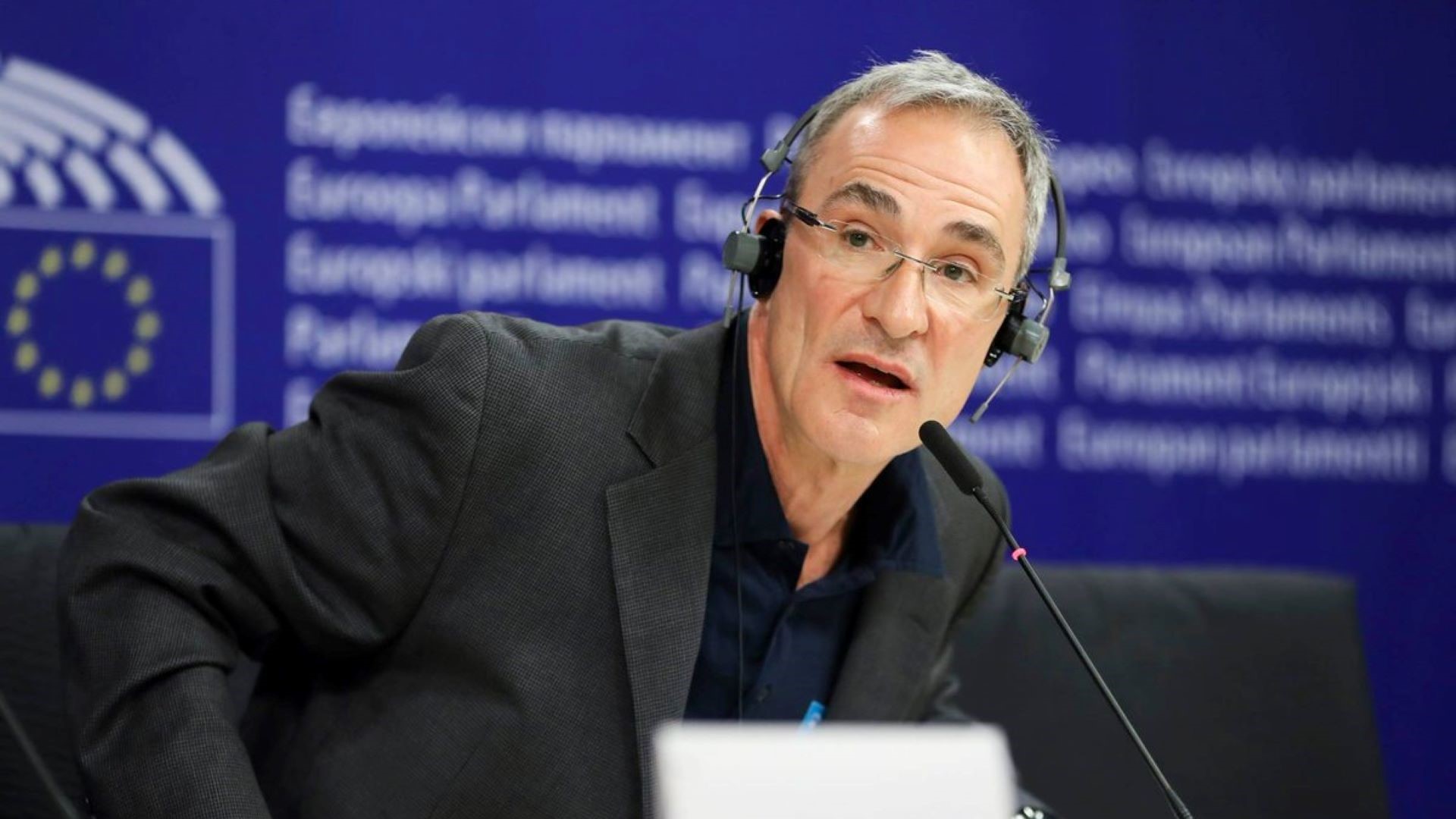 Head and shoulders photo of Dr Paul Dorfman with headset on, sat in front of a microphone on a desk, talking at an event with an EU logo on screen behind