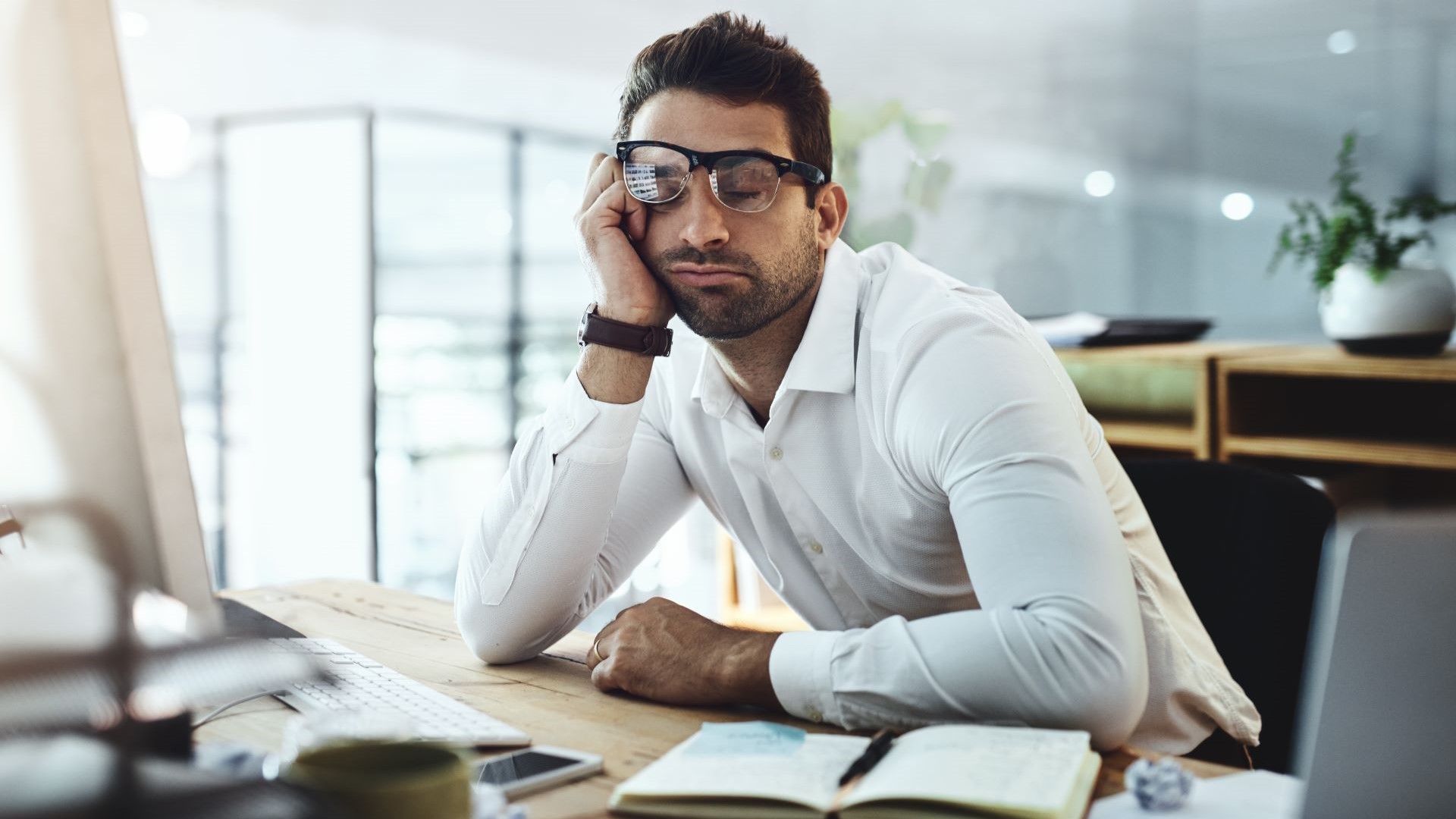 Man half asleep, sat at a work desk, elbows on table, one hand supporting face and knocking glasses askew