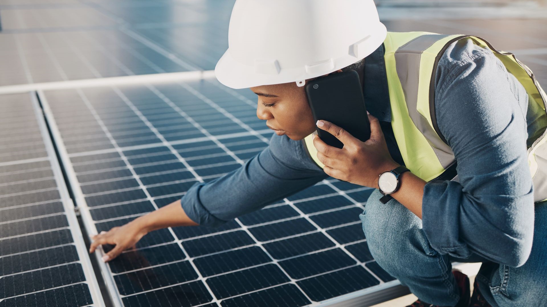 Woman wearing a hard hat and hi vis jacket using a phone and inspecting a solar panel