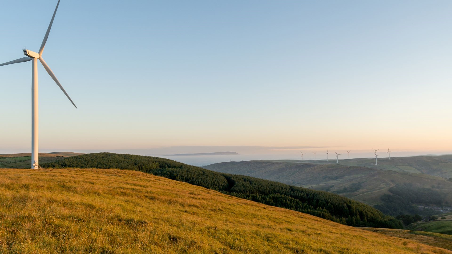 Single wind turbine on top of grassy hill, with hilly countryside and more wind turbines extending into distance behind 