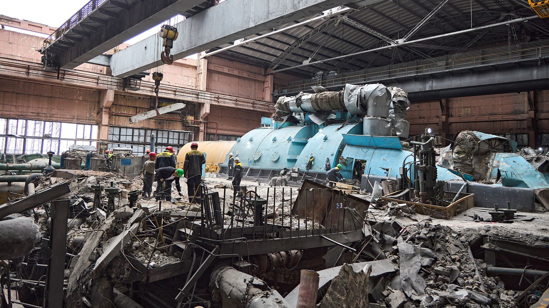 Wreckage of power plant destroyed by bomb, twisted pipework and metal, collapsed roof, and three workers clearing debris