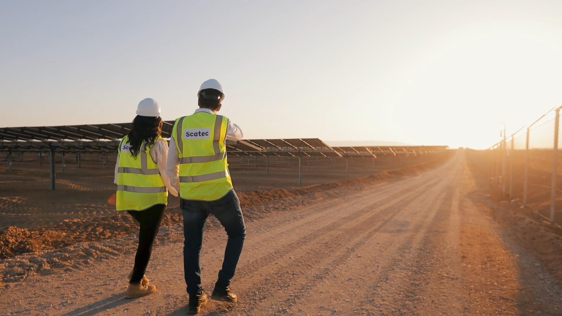 Two people in safety wear walking along a path near rows of solar panels