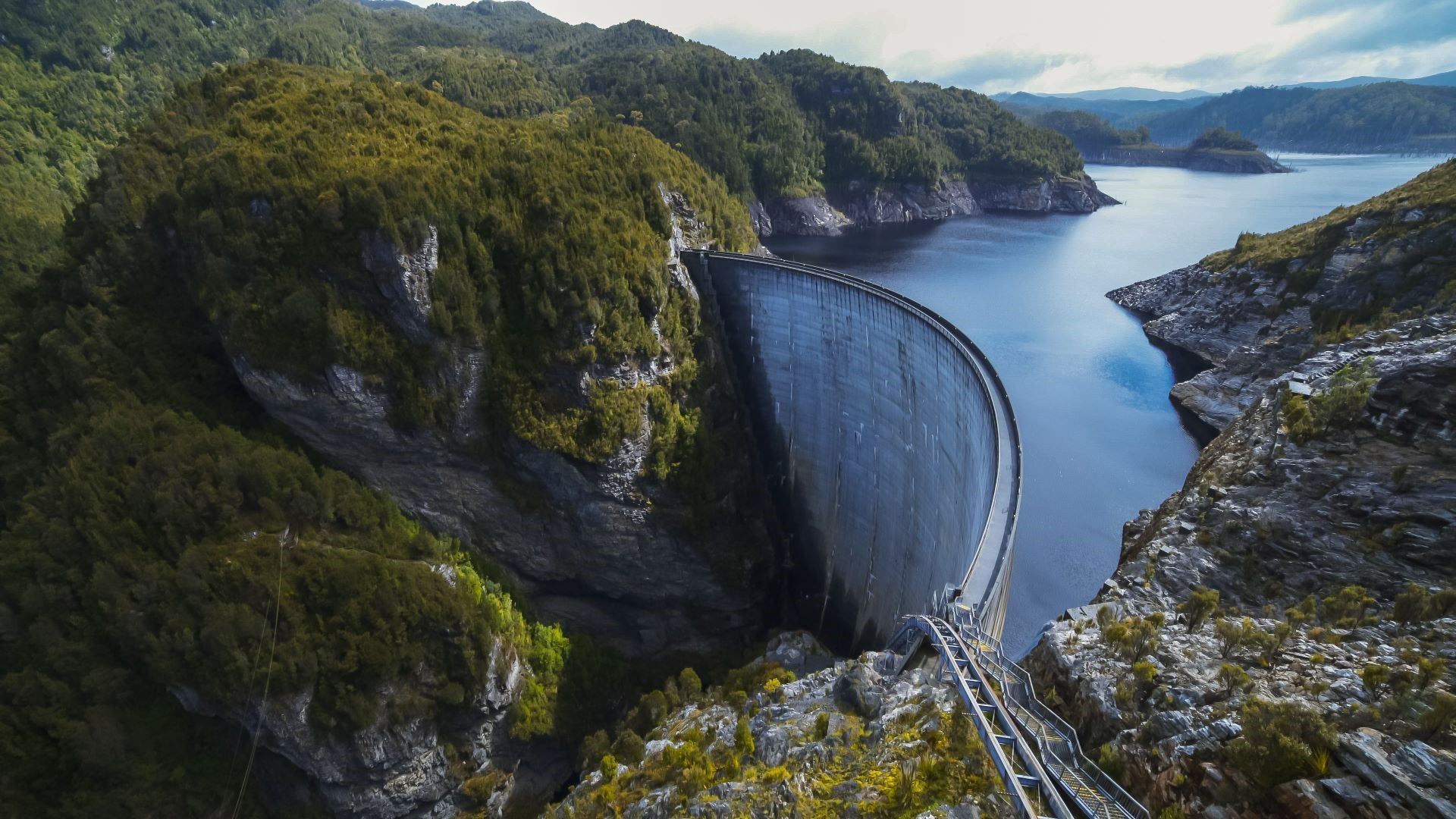 Arial overview of dam in mountainous region, with calm blue waters behind it, blue ski and clouds above