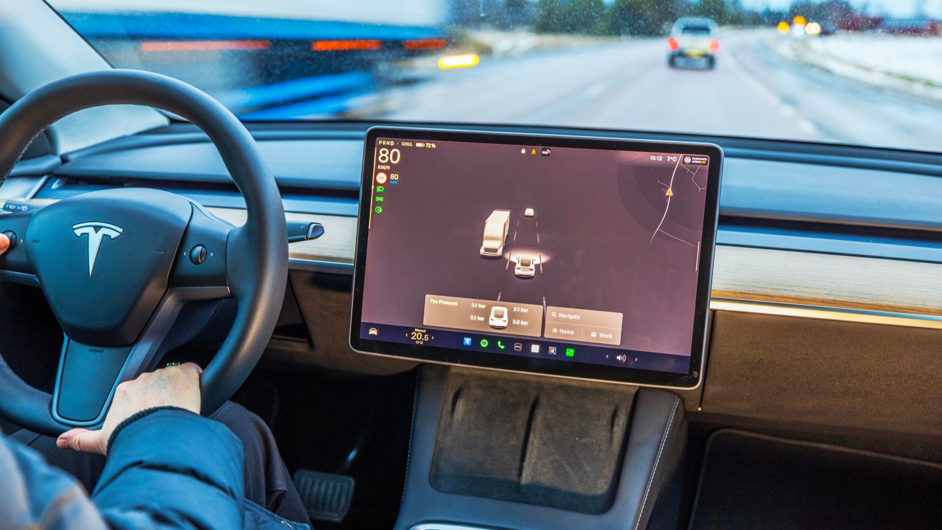 Close up of hands on an electric vehicle's steering wheel on left hand side of car, with computer screen to right of steering wheel, looking onto a road with a few other cars and a truck
