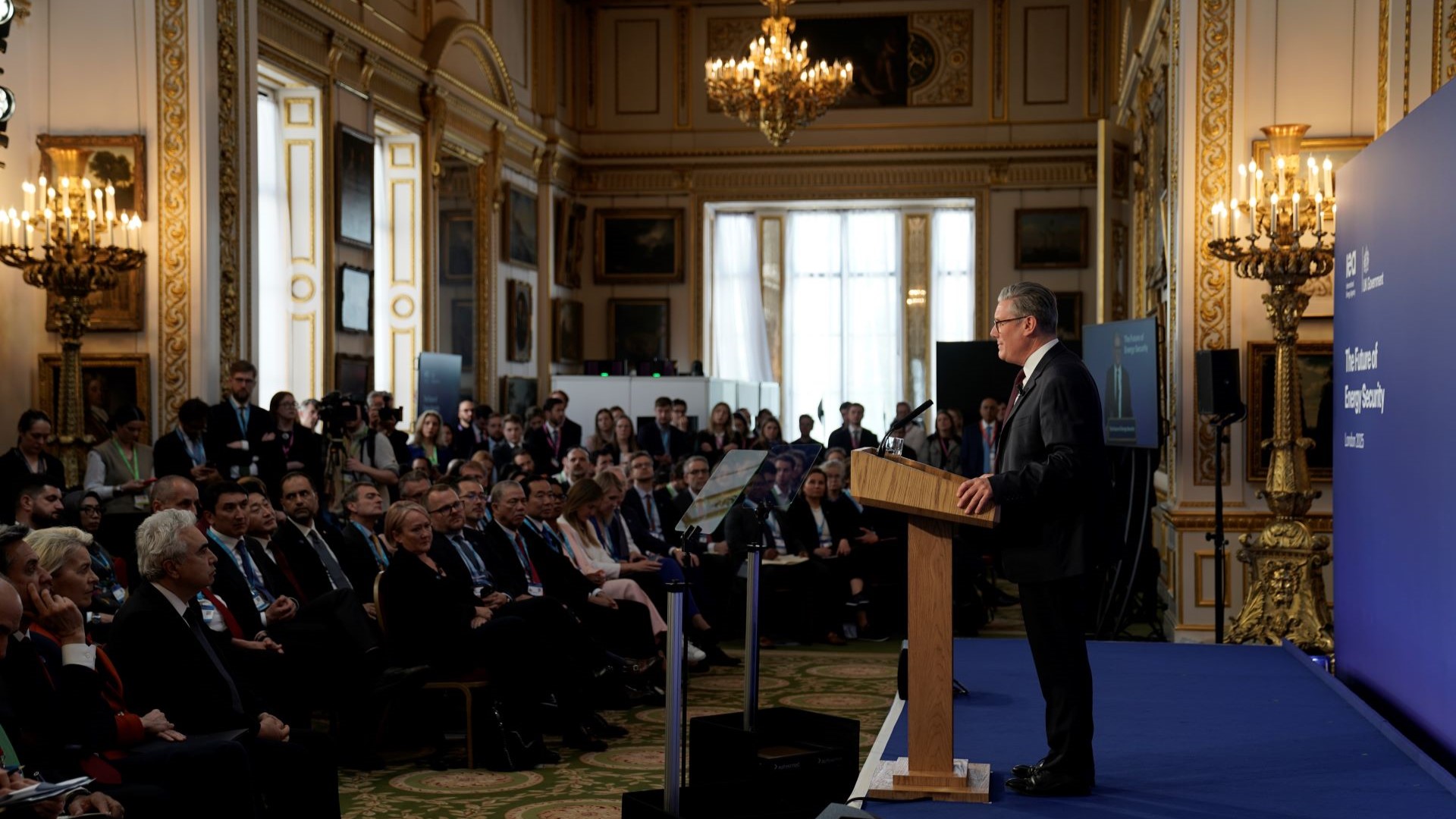 Grand room with glass chandelier, filled with men and women seated in rows, listening to man stood at lectern