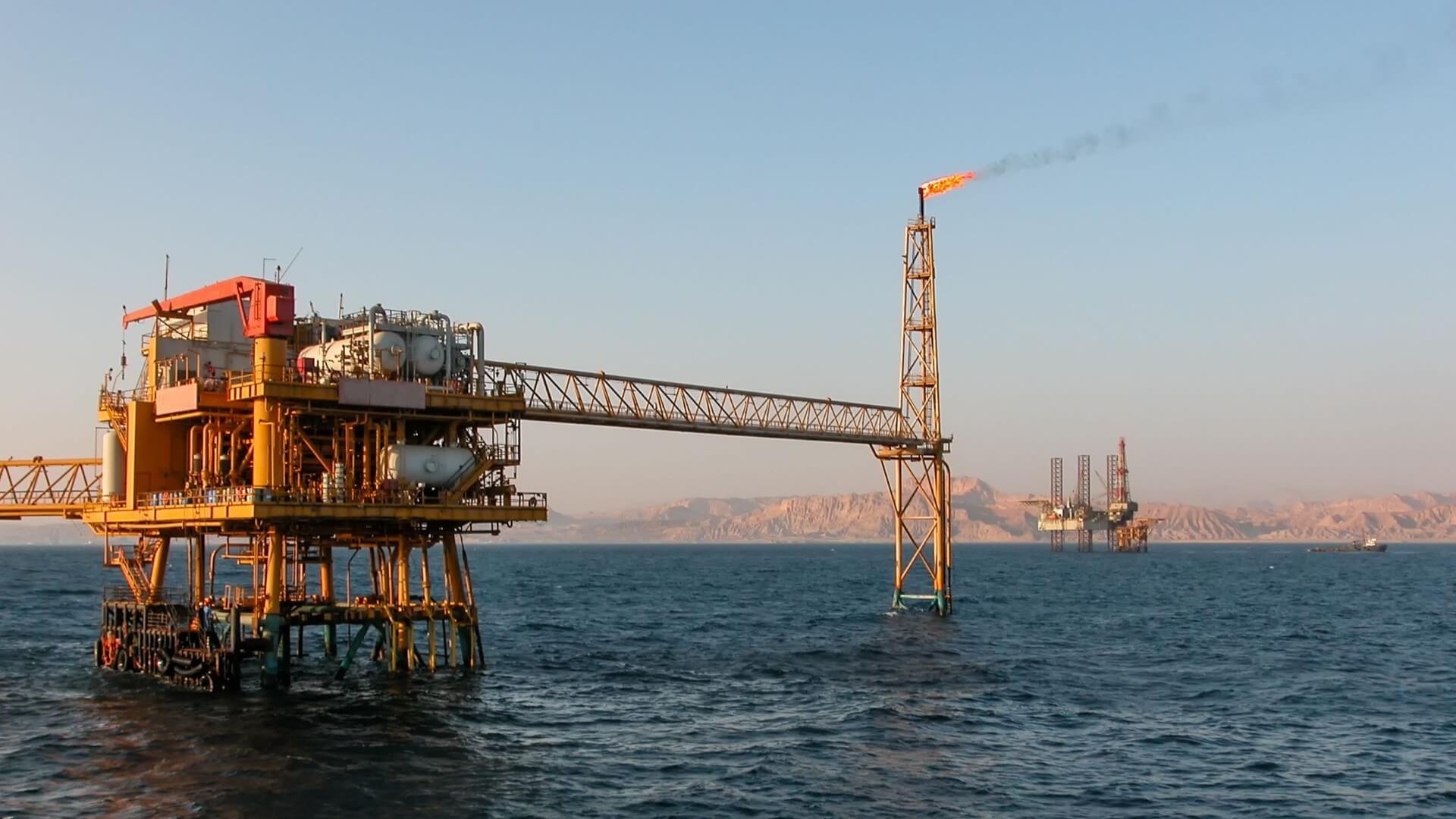 View from sea of oil production platform flaring gas from gas flare set to one side, with view of another platform and land in distance