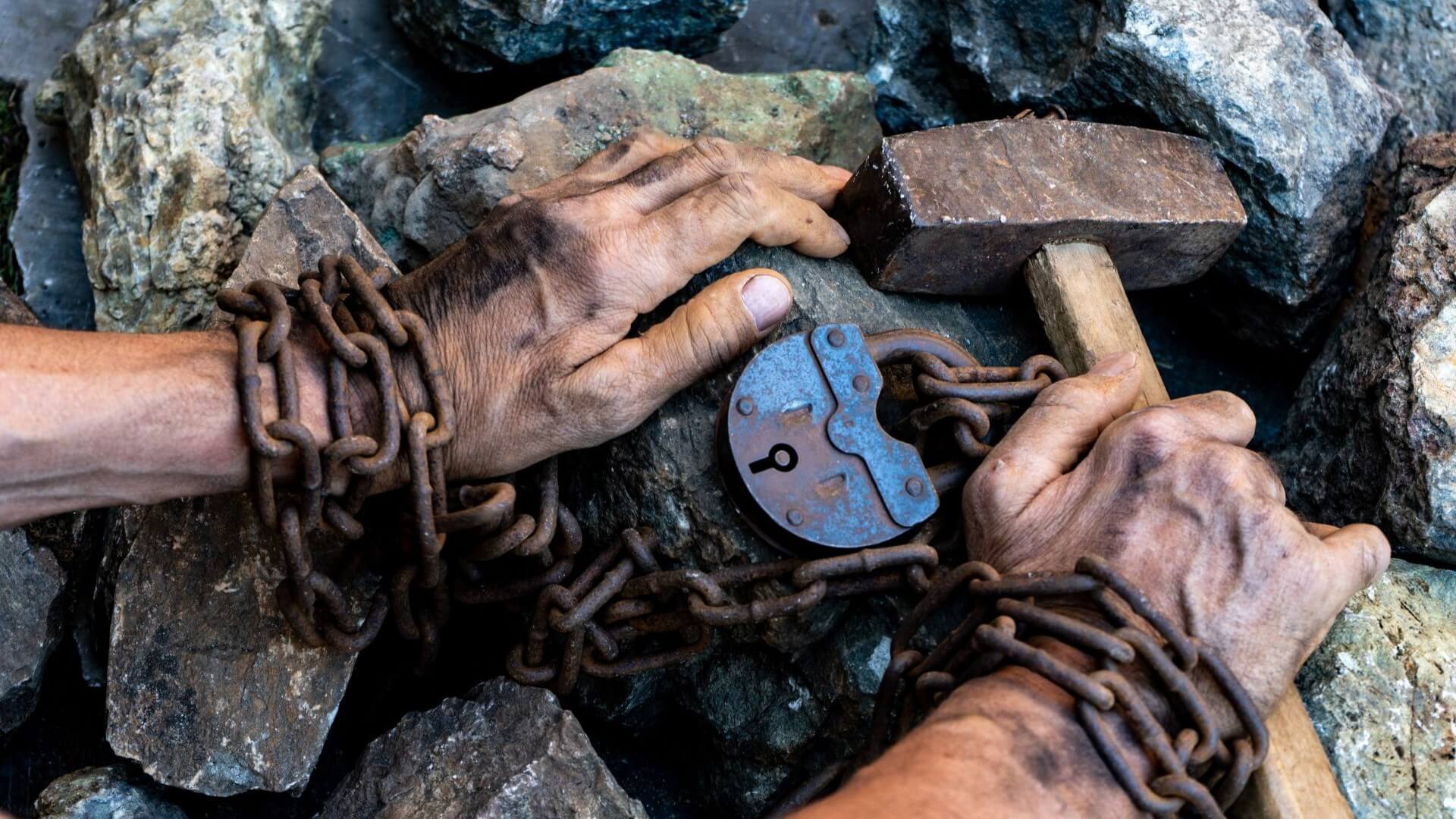 Close up of hands manacled with with chain and padlock, resting on a pile of hand sized rocks, with one hand holding a large hammer to break up the rocks