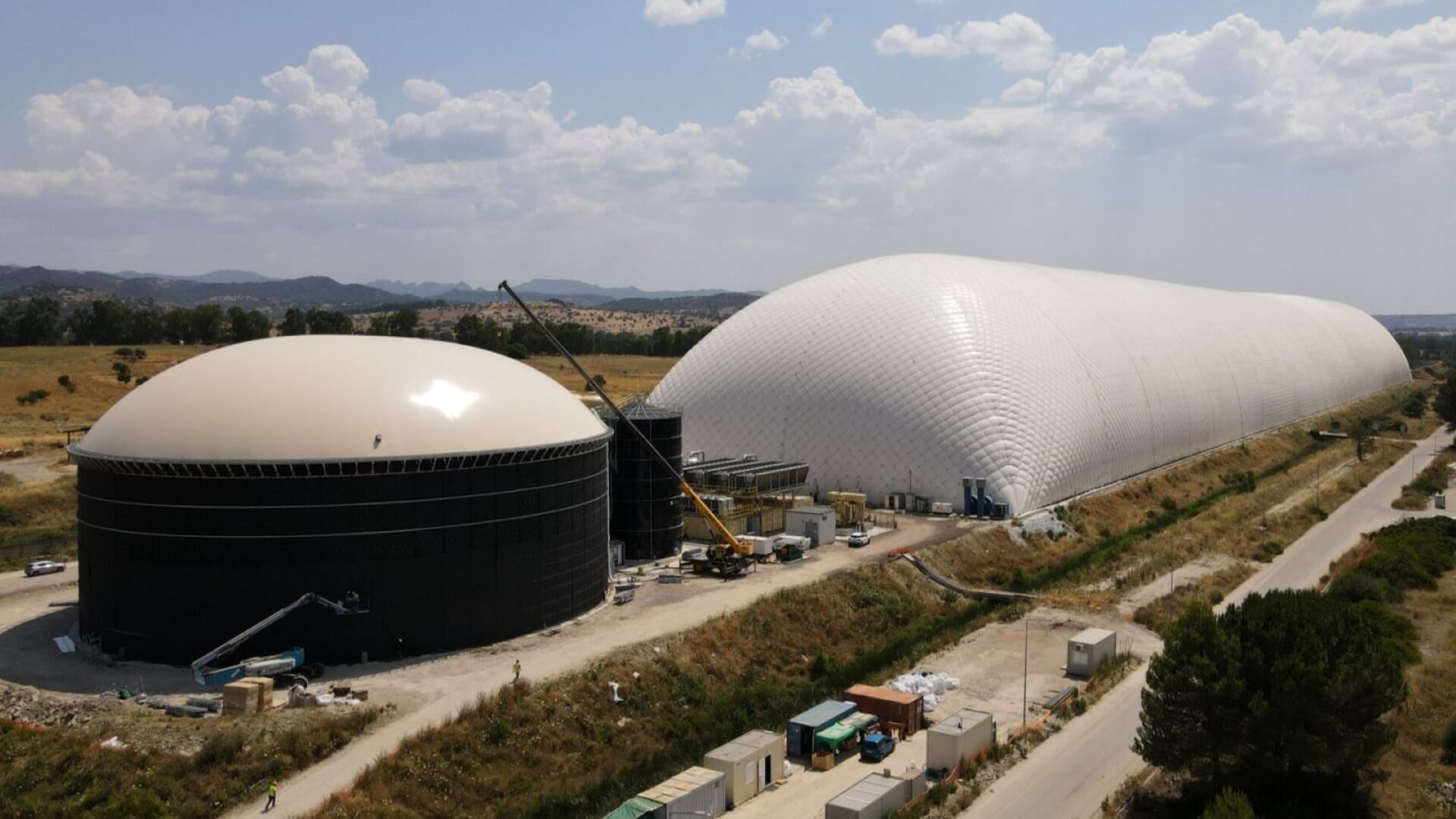 Aerial view of large circular domed metal storage tank to left, with long rectanglur 'flexible' sided dome shaped 'gas bag' to right; road and cars parked nearby