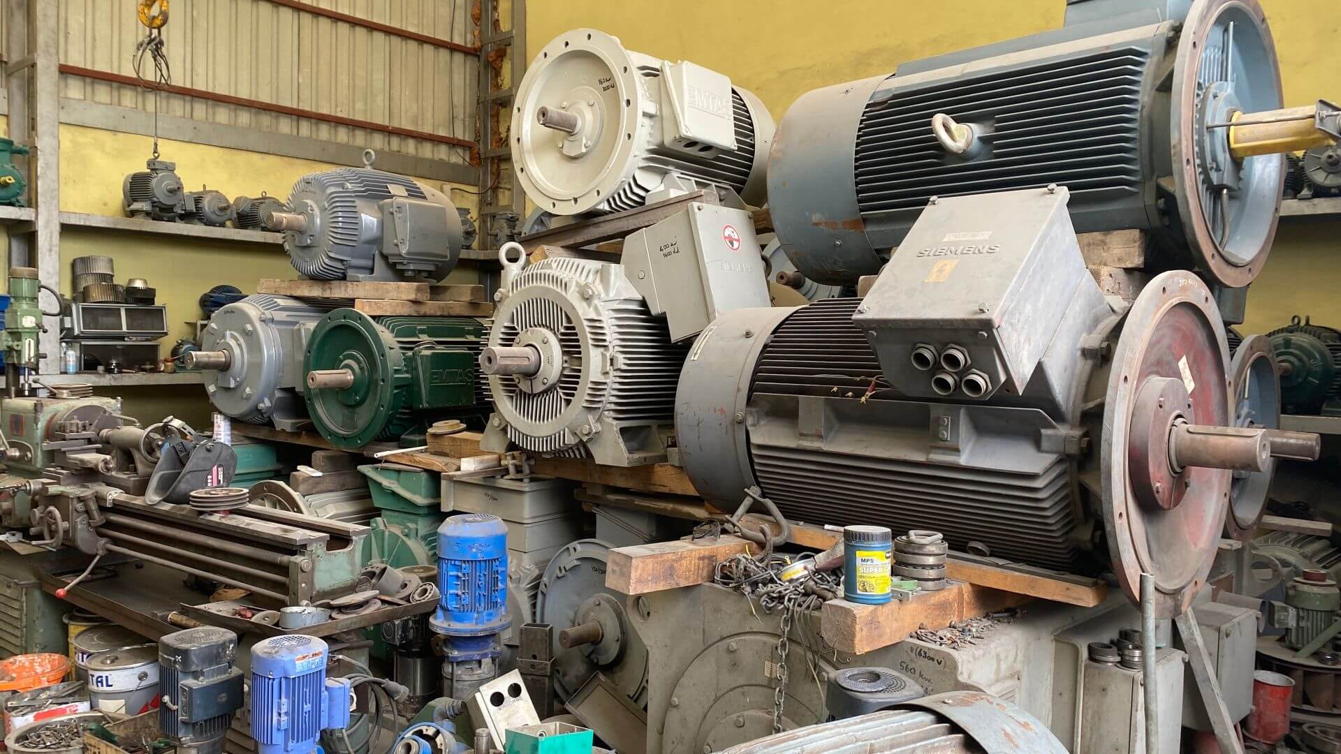 Large metal motor with rotating cylinders on a bench in a workshop, surrounded by other bits of old equipment waiting to be recycled
